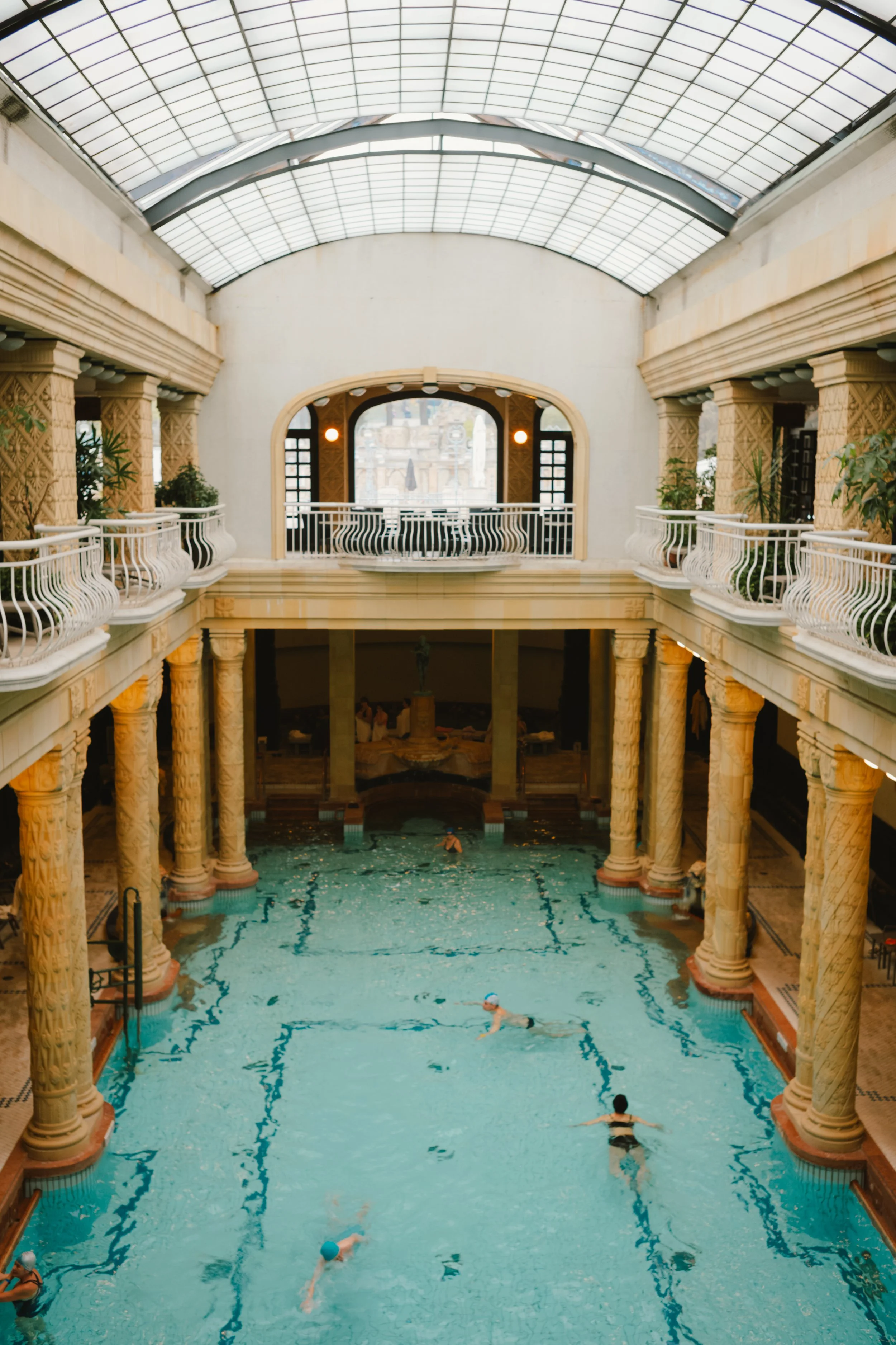 Indoor swimming pool with swimmers, surrounded by columns and balcony, with a domed glass ceiling letting in natural light.