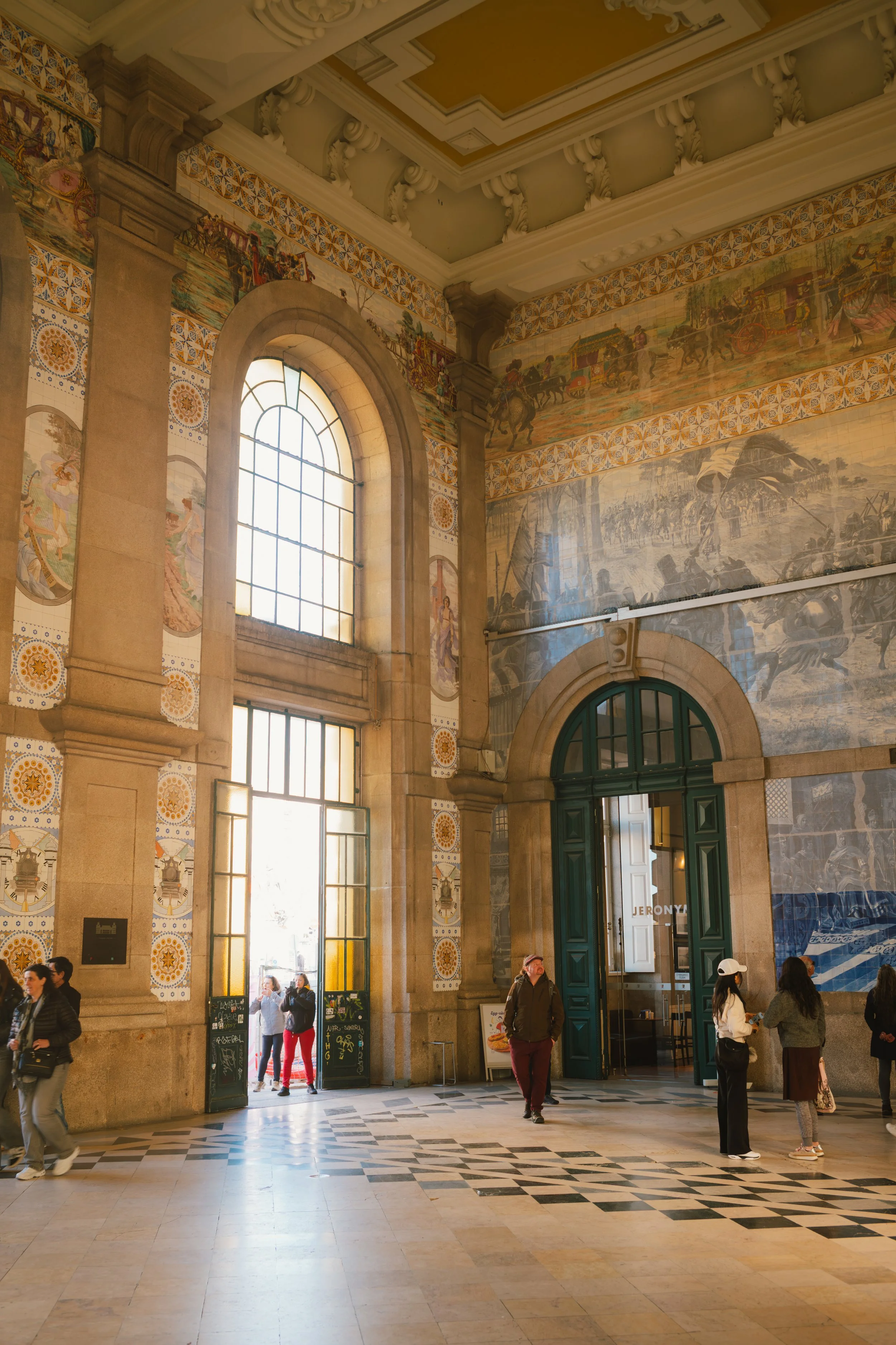 Interior of a historic train station with large arched windows, ornate tilework, and wall murals depicting historical scenes. Several people are walking or standing near the entrance.