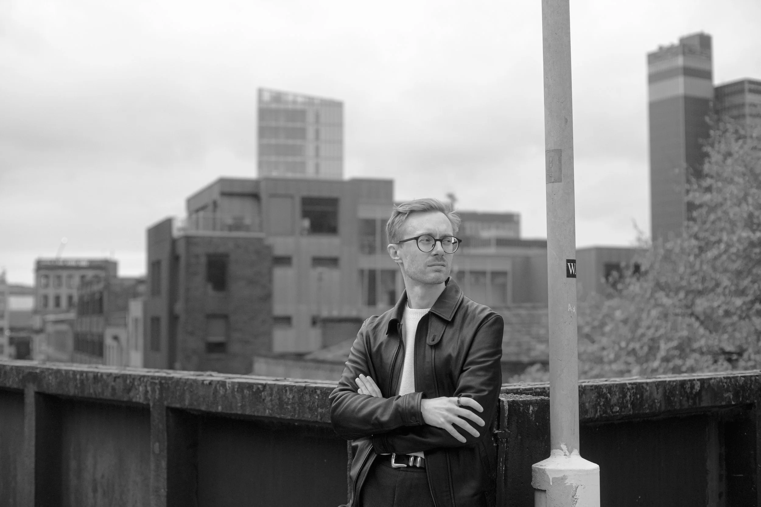 A man with glasses and a jacket stands outdoors in an urban area with modern buildings in the background. He is crossing his arms and looking to the side near a streetlight.
