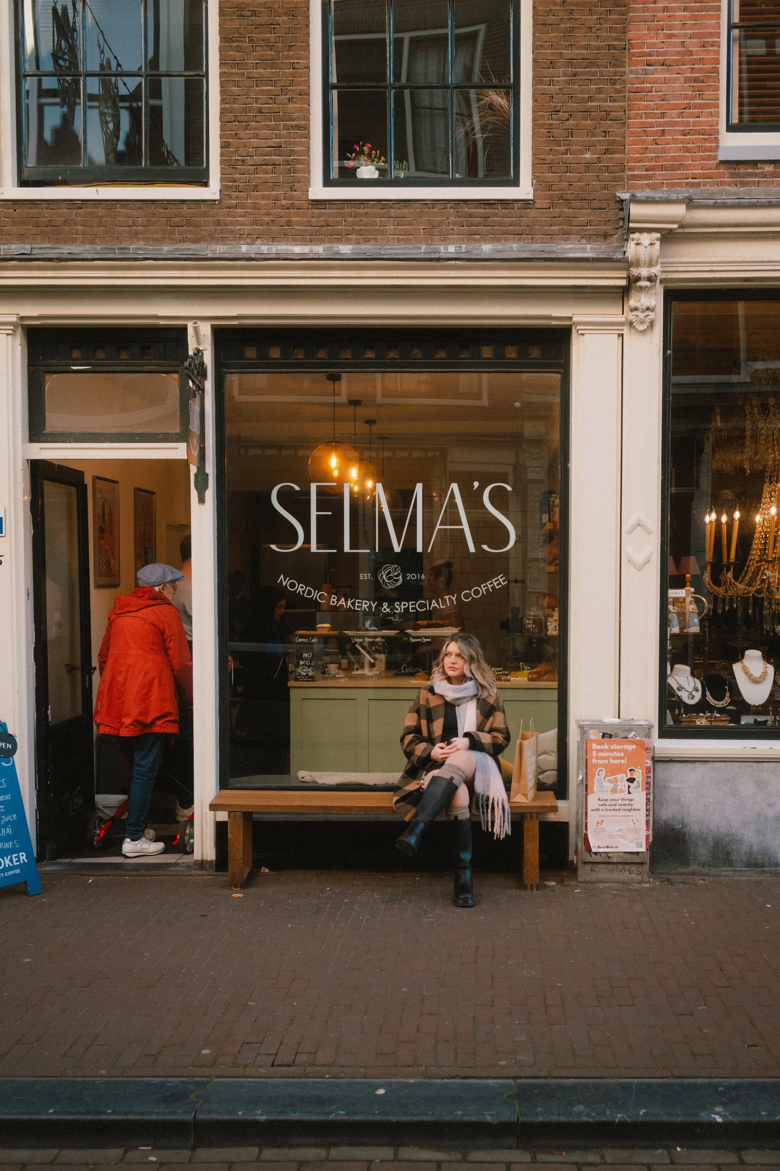 Street view of a cafe named 'SELMAS' with a large window and an open door. Inside, people are sitting and dining, and a woman is sitting on a wooden bench outside, wearing a coat, scarf, and black boots. There is a woman in an orange coat and gray ha