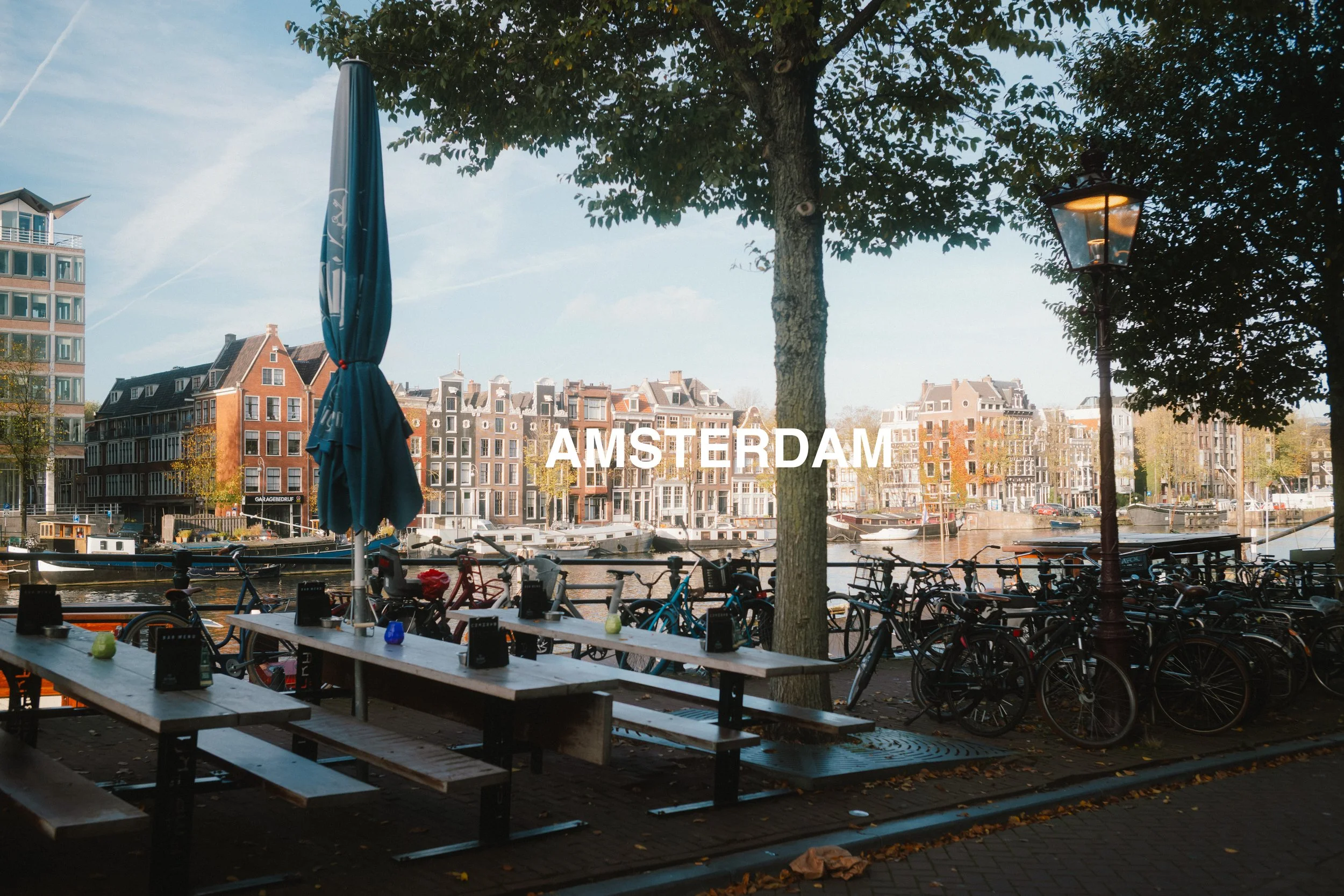 View of a canal in Amsterdam with historic buildings, bicycles parked along the sidewalk, a picnic table, and a tree with a street lamp, under a partly cloudy sky.