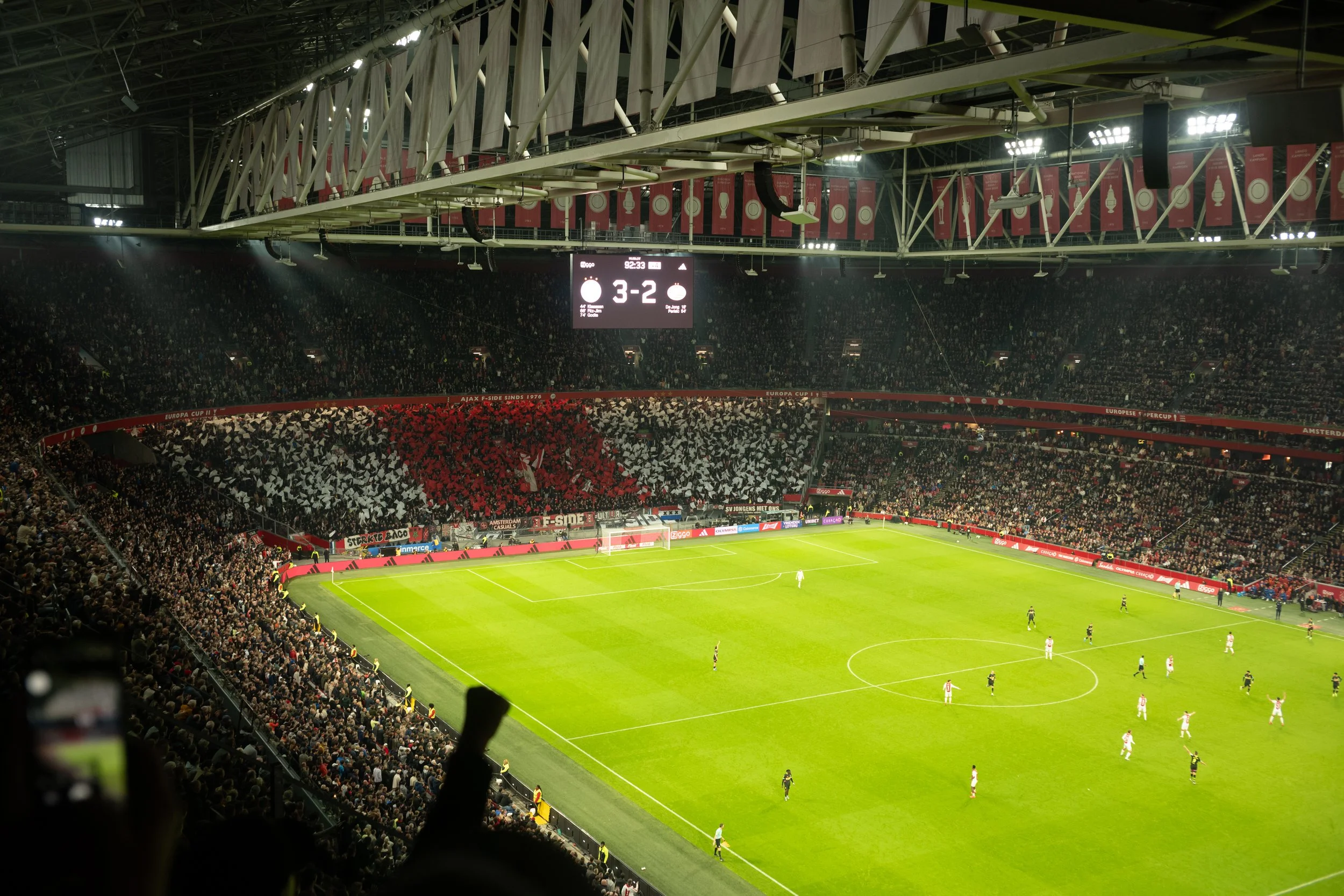 Soccer stadium filled with spectators watching a game. The scoreboard shows a score of 3-2. The field is green with players visible.