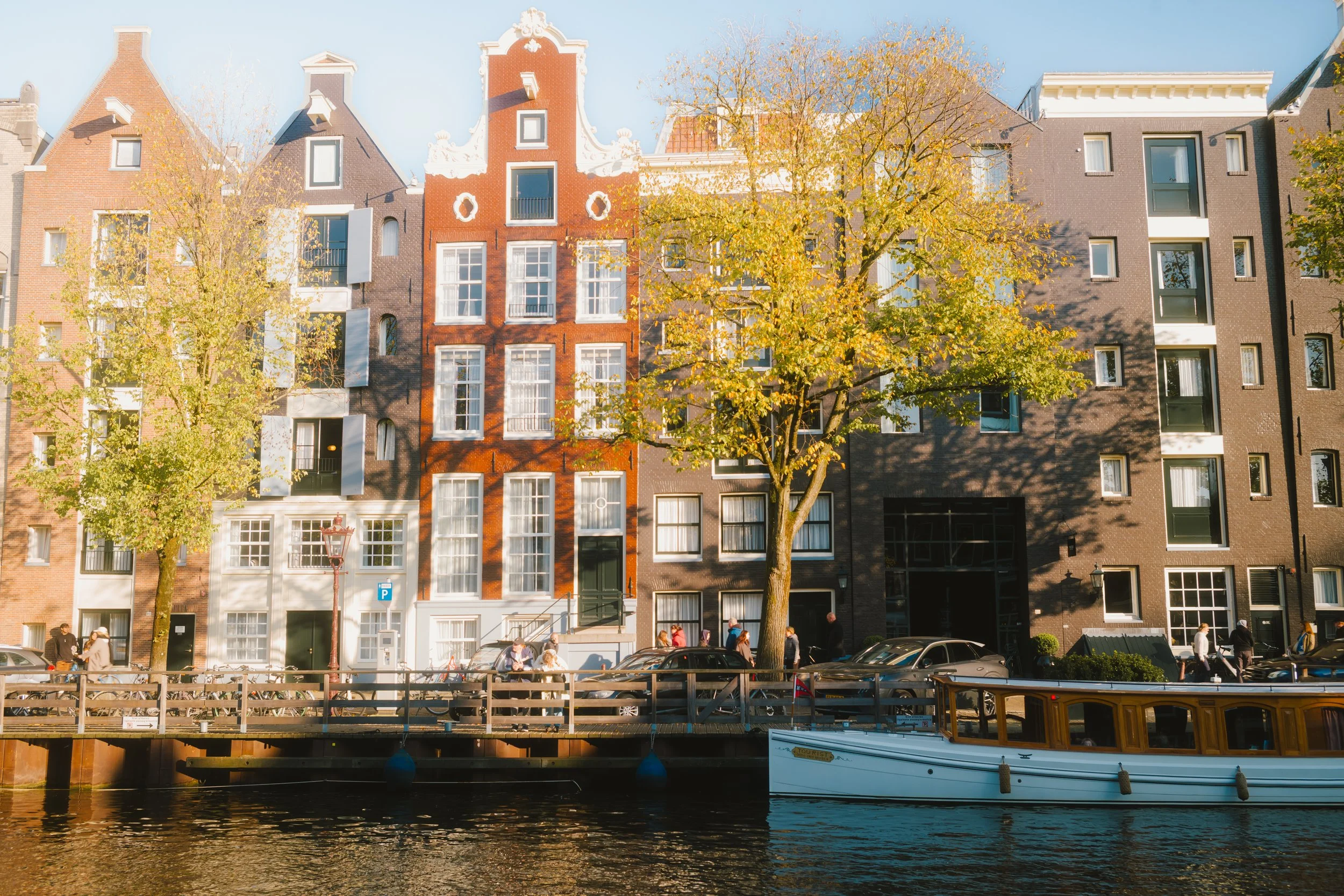 Colorful row of historic buildings along a canal, with trees showing fall foliage, and a boat floating on the water in the foreground.