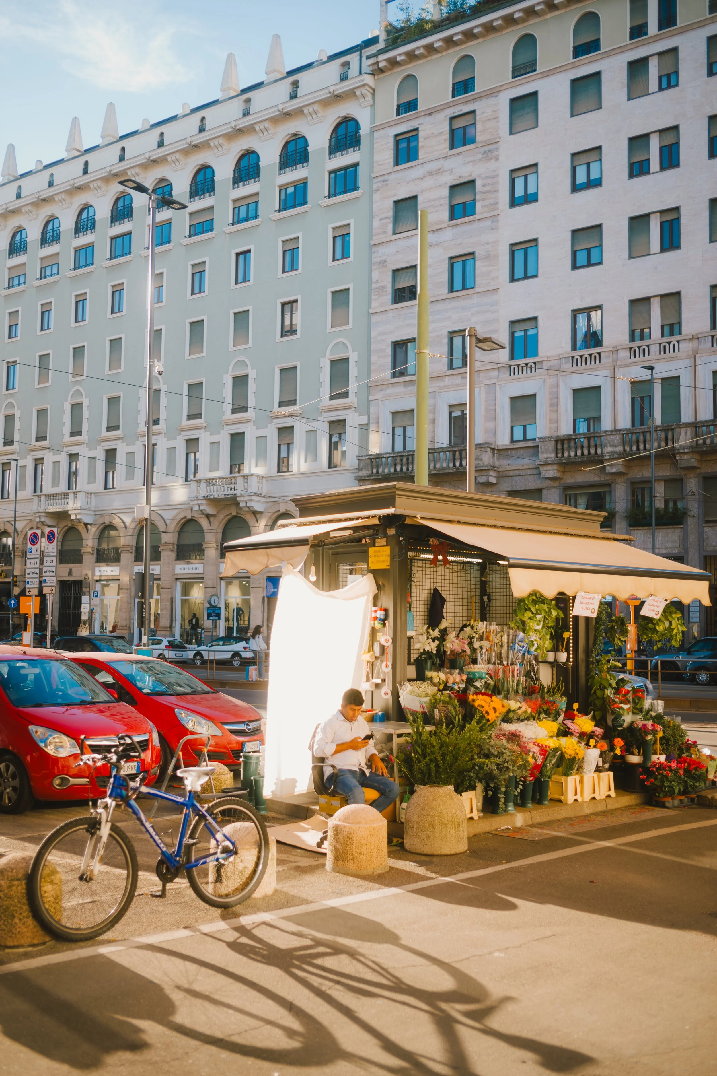 A street scene with a flower vendor stand, a man sitting with a phone, several parked cars including red ones, bicycles, and a tall building with multiple windows and ornate architectural details in the background, during daylight.