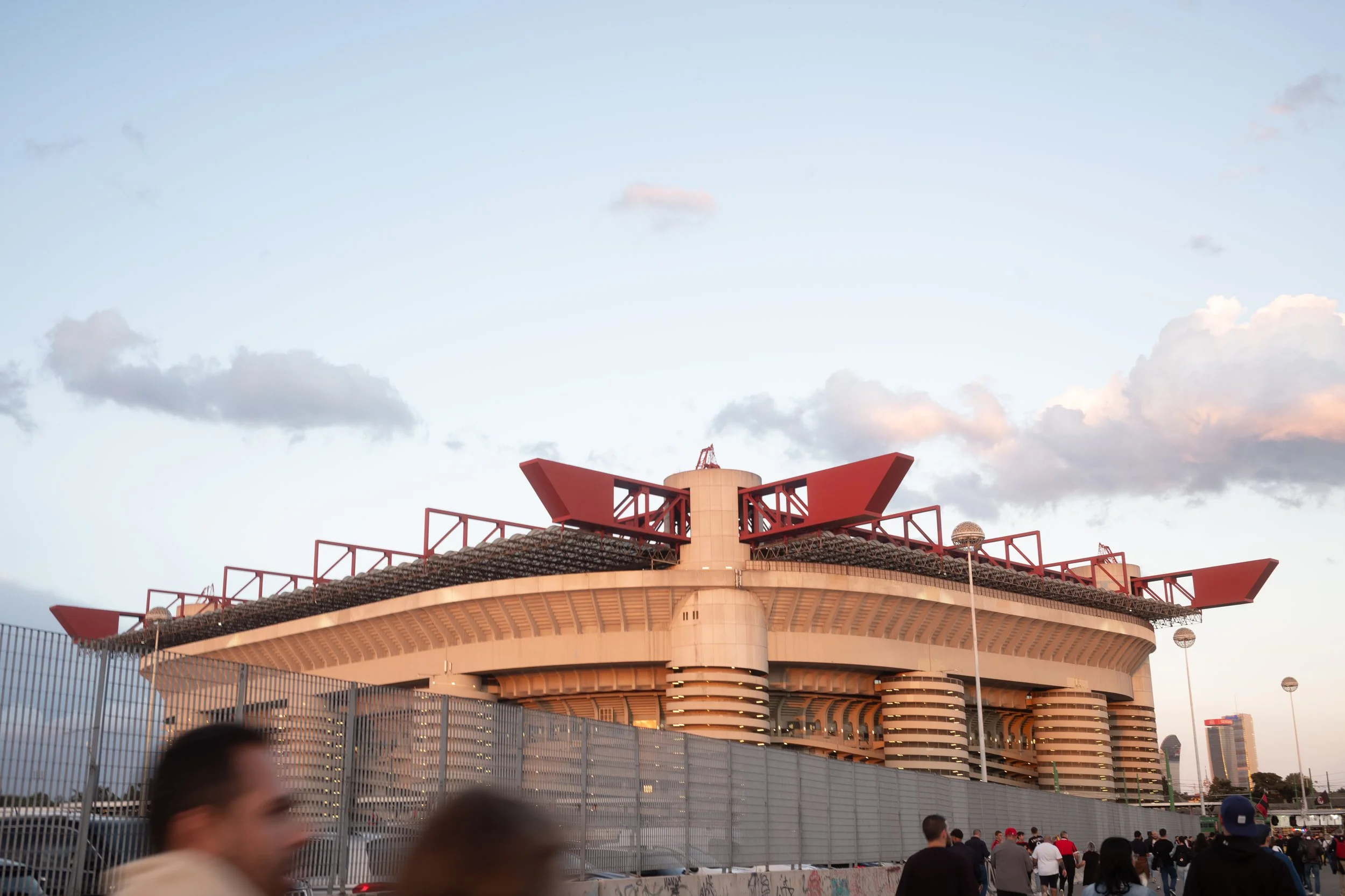 A large circular building with a red architectural structure on top, surrounded by a metal fence and a crowd of people in the foreground, under a partly cloudy sky.