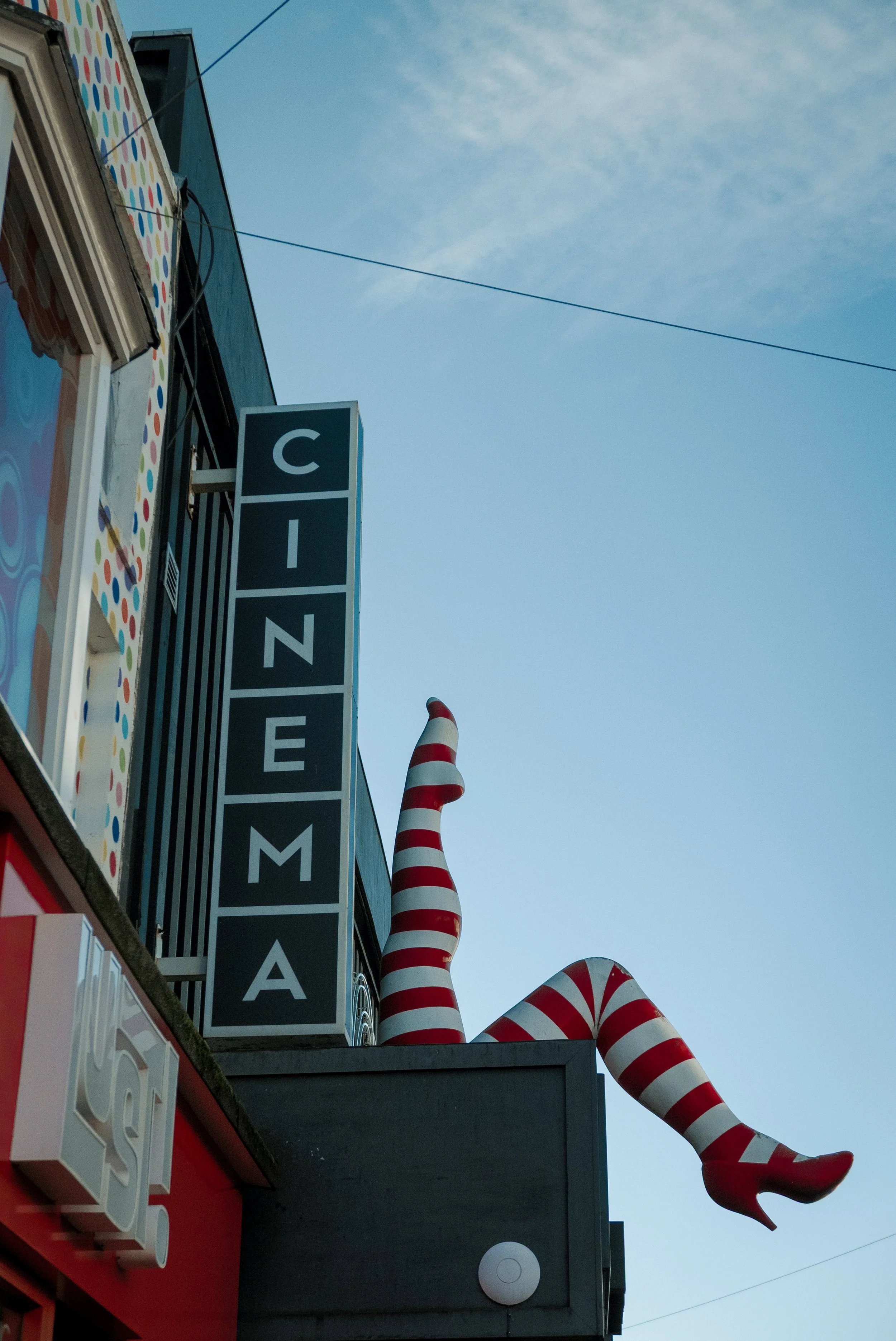 Photo of a building with a sign that reads 'CINEMA'. There are three large red and white striped carnival-themed lollipops or candy canes hanging from the building's wall. The sky is clear with some clouds.