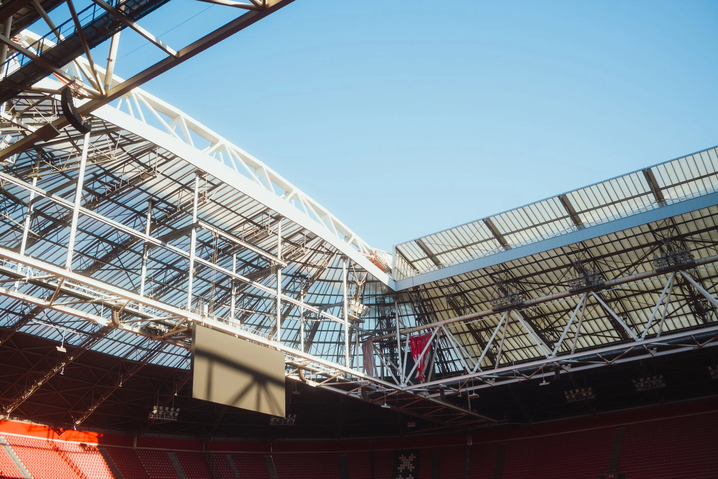A view of the interior of a stadium under construction with a large steel framework, glass roof, and red seating area.