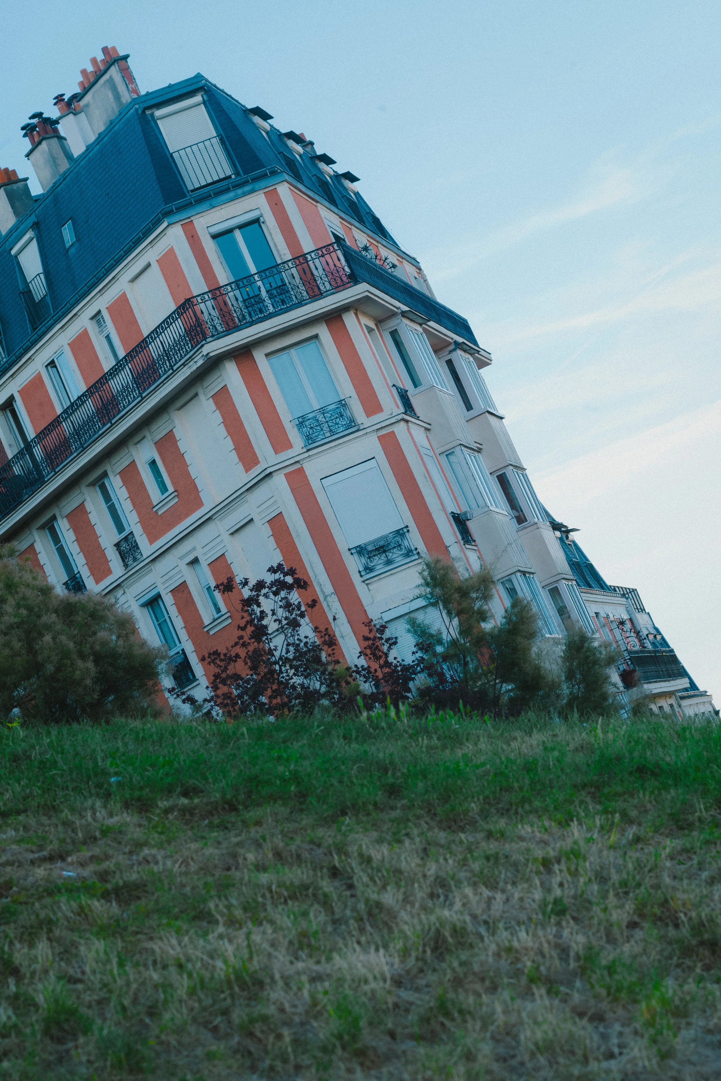 A multi-story residential building with colorful exterior walls and black wrought-iron balconies, seen from a low angle with grass and bushes in the foreground and a clear blue sky above.