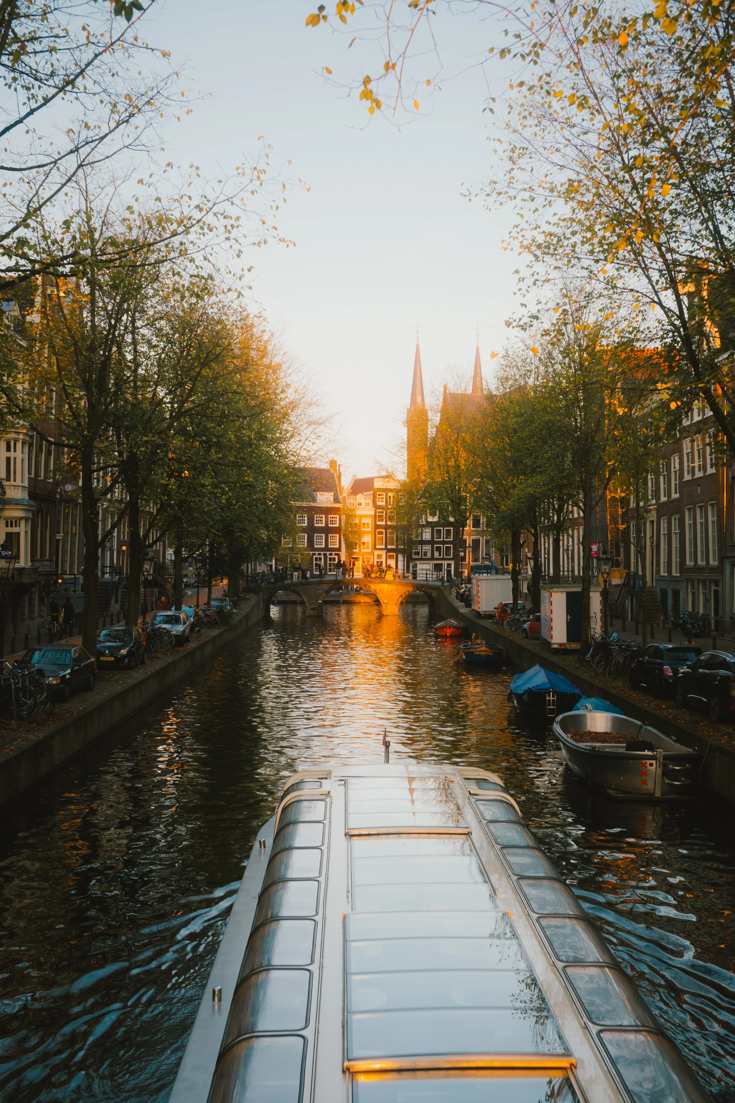 A canal with boats docked along the sides, trees with autumn leaves, and a church with twin spires in the background during sunset.