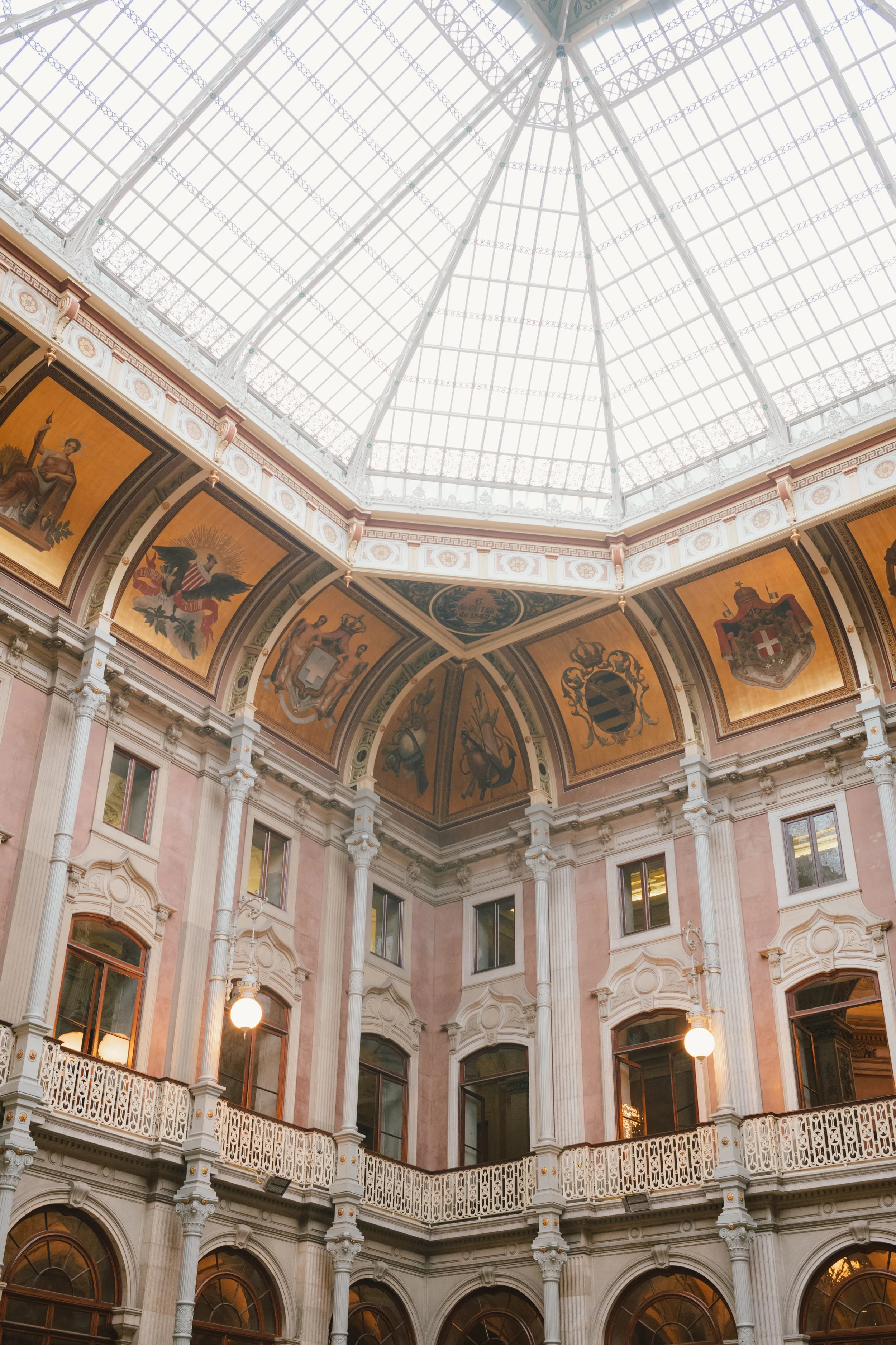 Interior view of a grand historic building with a large glass dome ceiling, ornate architectural details, murals, arches, decorative columns, and warm lighting.