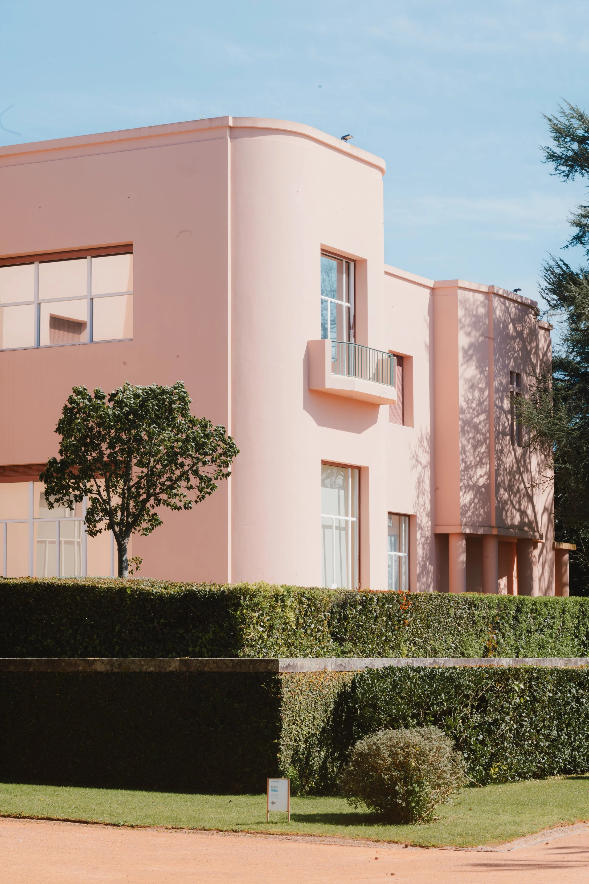 A pink, modern-style apartment building with rectangular windows, a small balcony, surrounded by greenery and trees, under a blue sky.