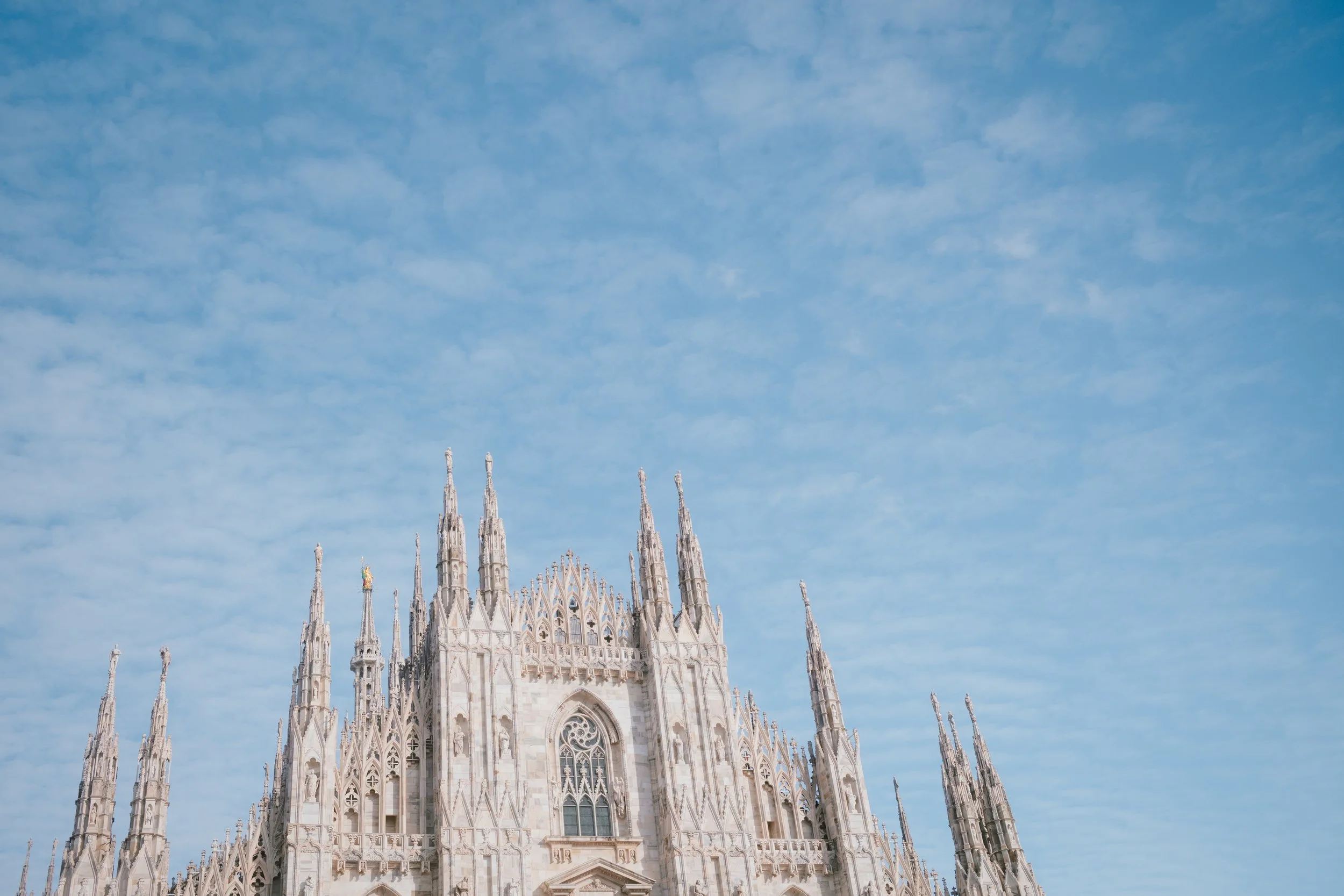 The top part of a historic cathedral with intricate architecture, pointed spires, and large arched windows, set against a bright blue sky with wispy clouds.