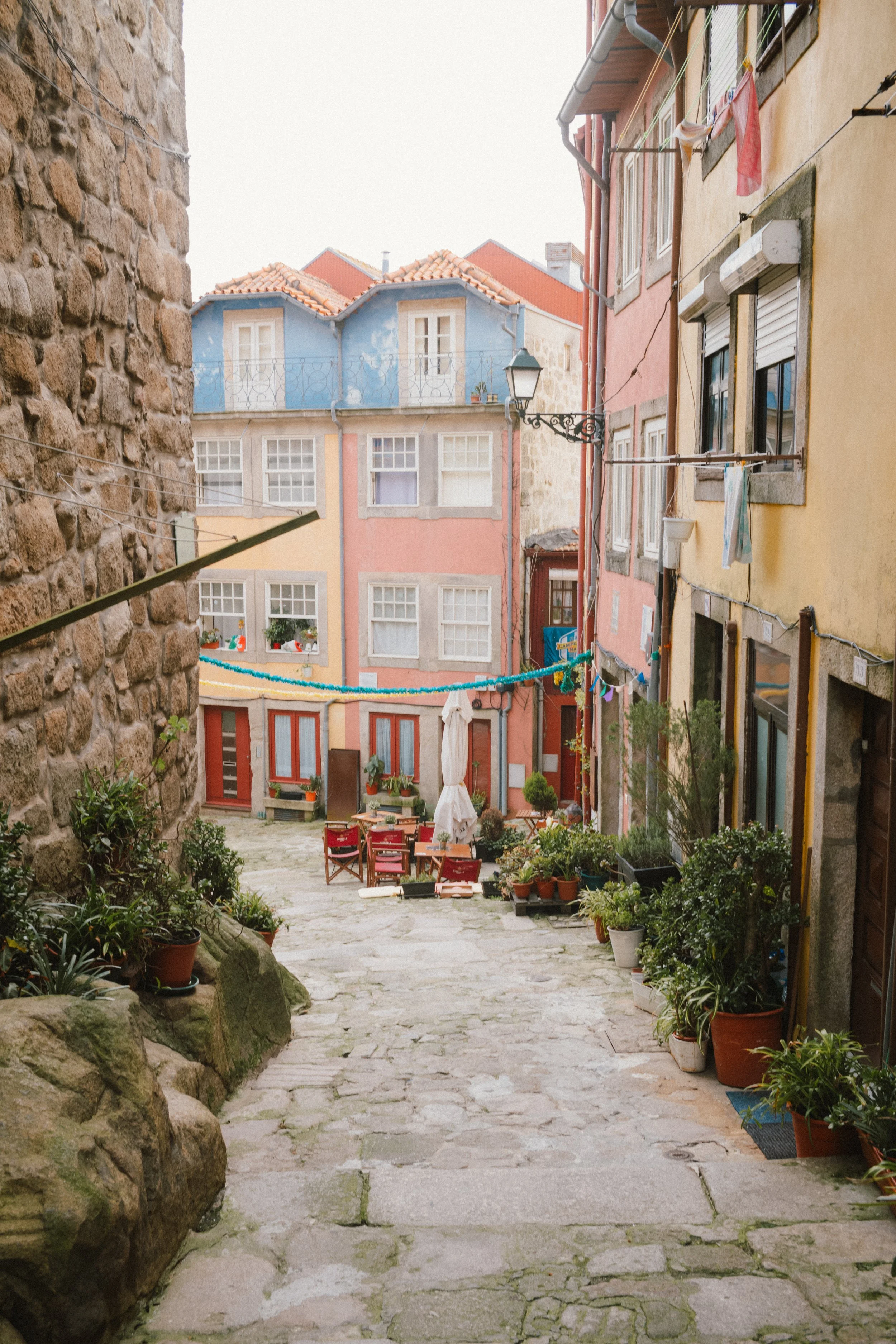 A European courtyard with cobblestone pavement, colorful buildings with red and blue facades, and potted plants lining the edges. There is outdoor dining furniture and laundry hanging on clotheslines between buildings.