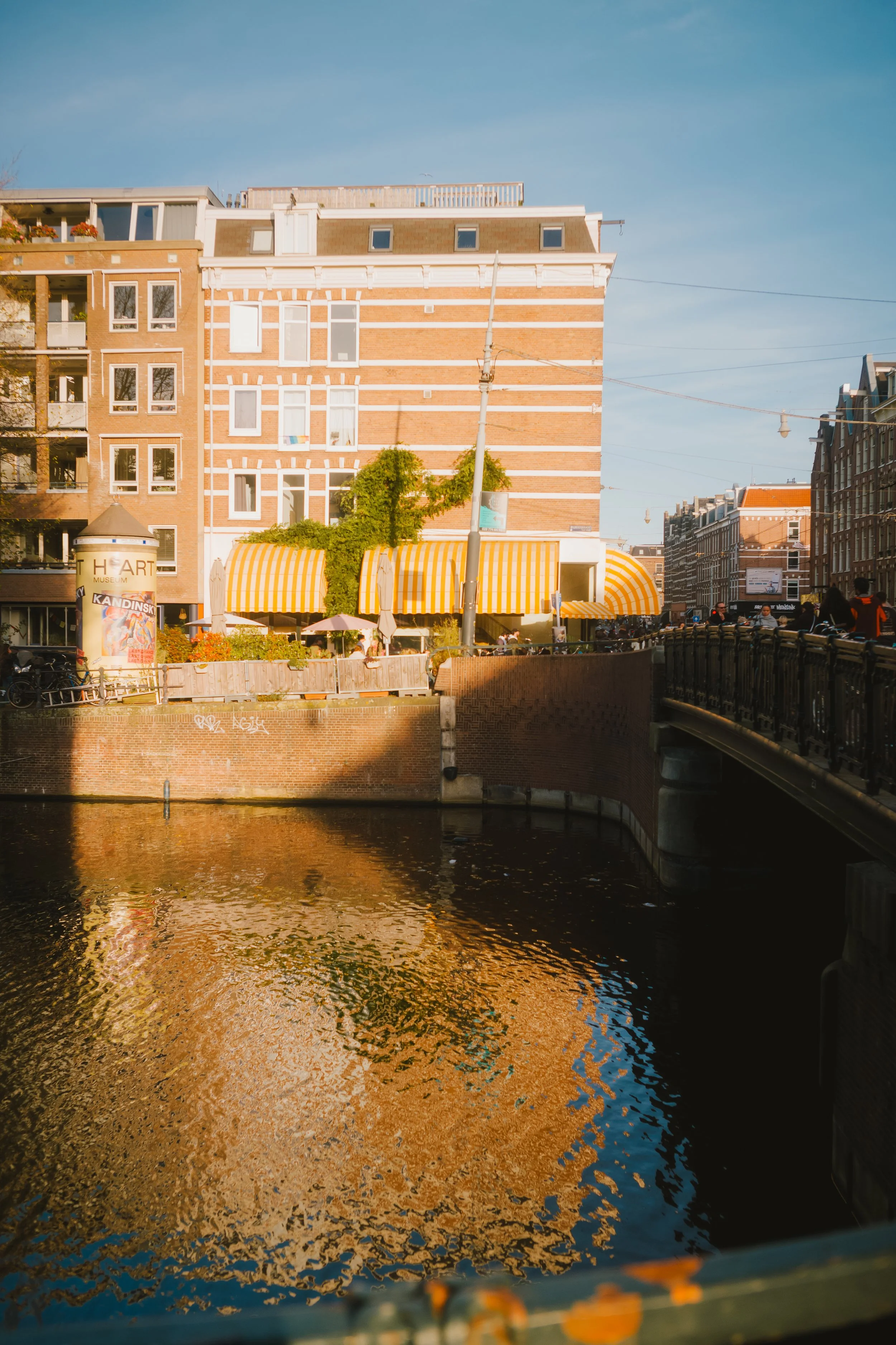 A cityscape featuring a river in the foreground, a bridge on the right, and a multi-story brick building with orange and white striped awnings and trees on the building's lower levels. The sky is clear with a few clouds.