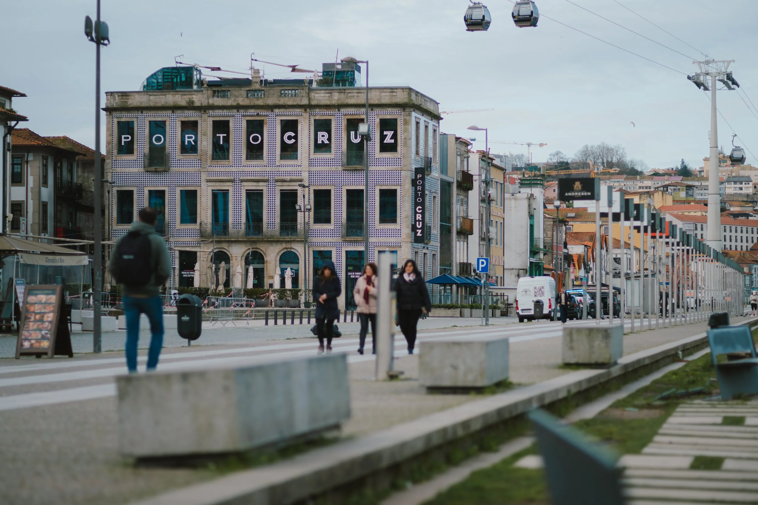 Public square in Portugal with a building displaying 'Porto Cruz' signage, people walking, and cable cars in the background.