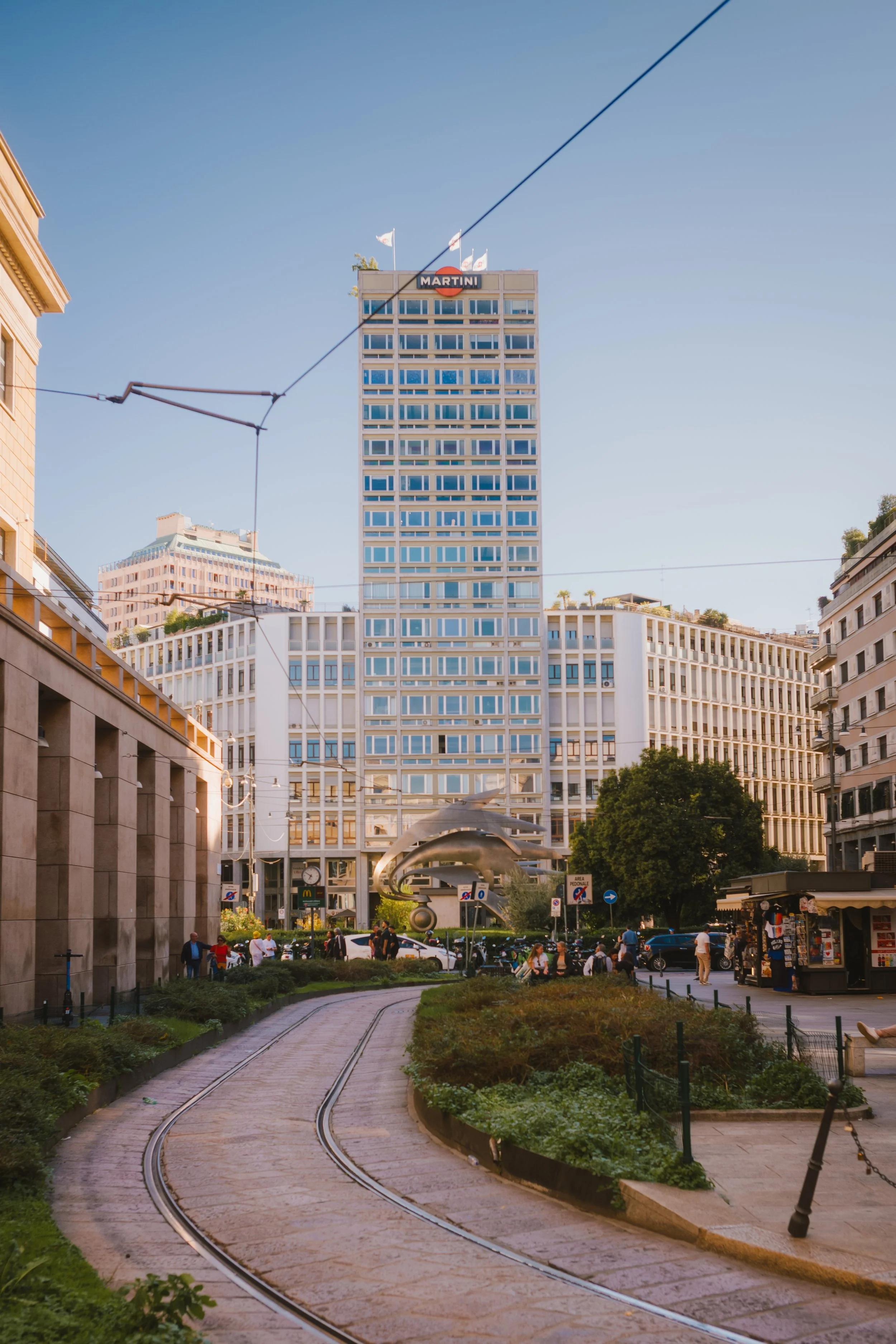 An urban scene with a curved tram track in the foreground, surrounded by green plants, and a busy pedestrian area with people walking and sitting. In the background, there are modern office buildings including a tall glass building with the sign 'Mar
