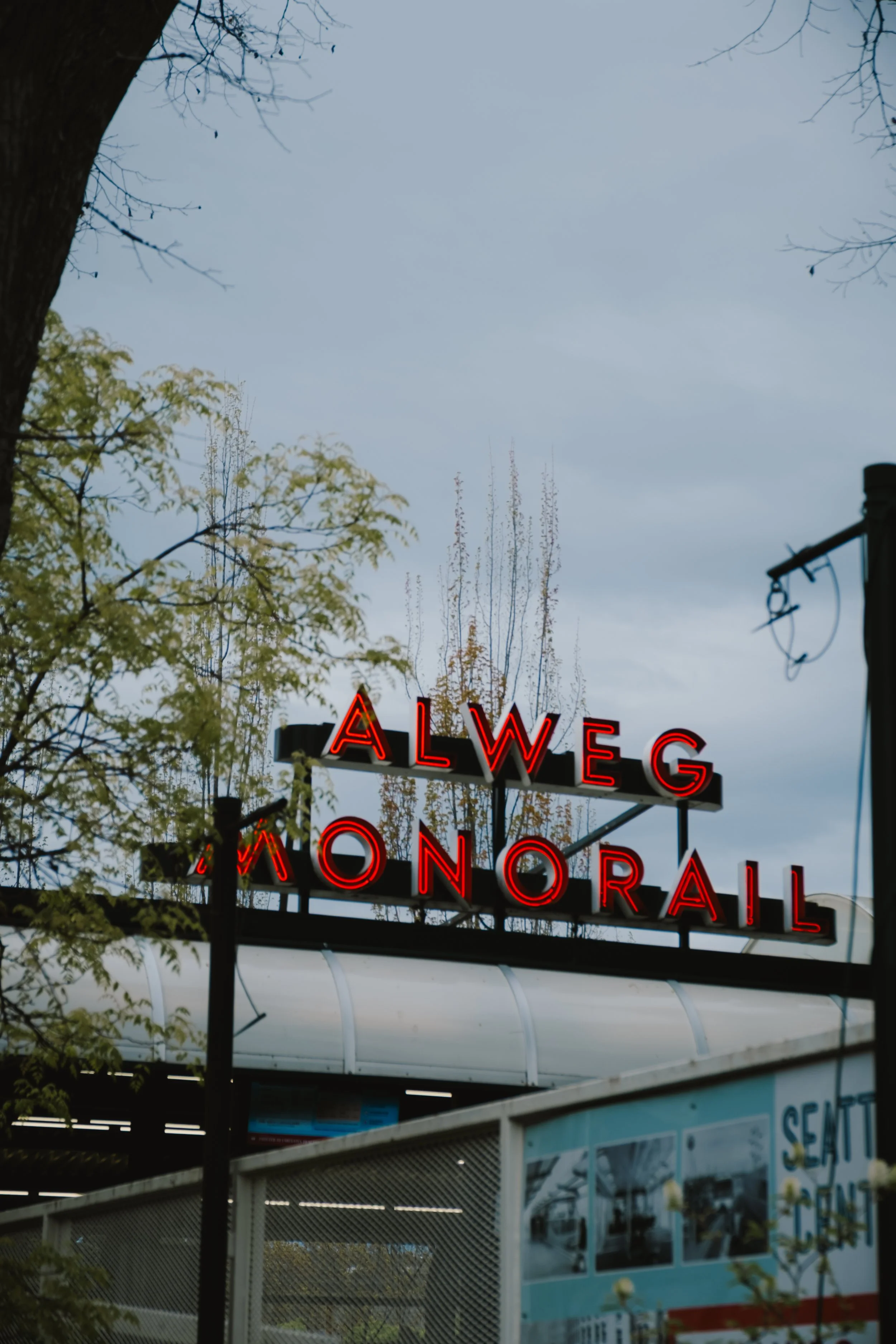 A partial view of a neon sign that reads 'ALWEG' and 'ONORAIL' with trees and a cloudy sky in the background.