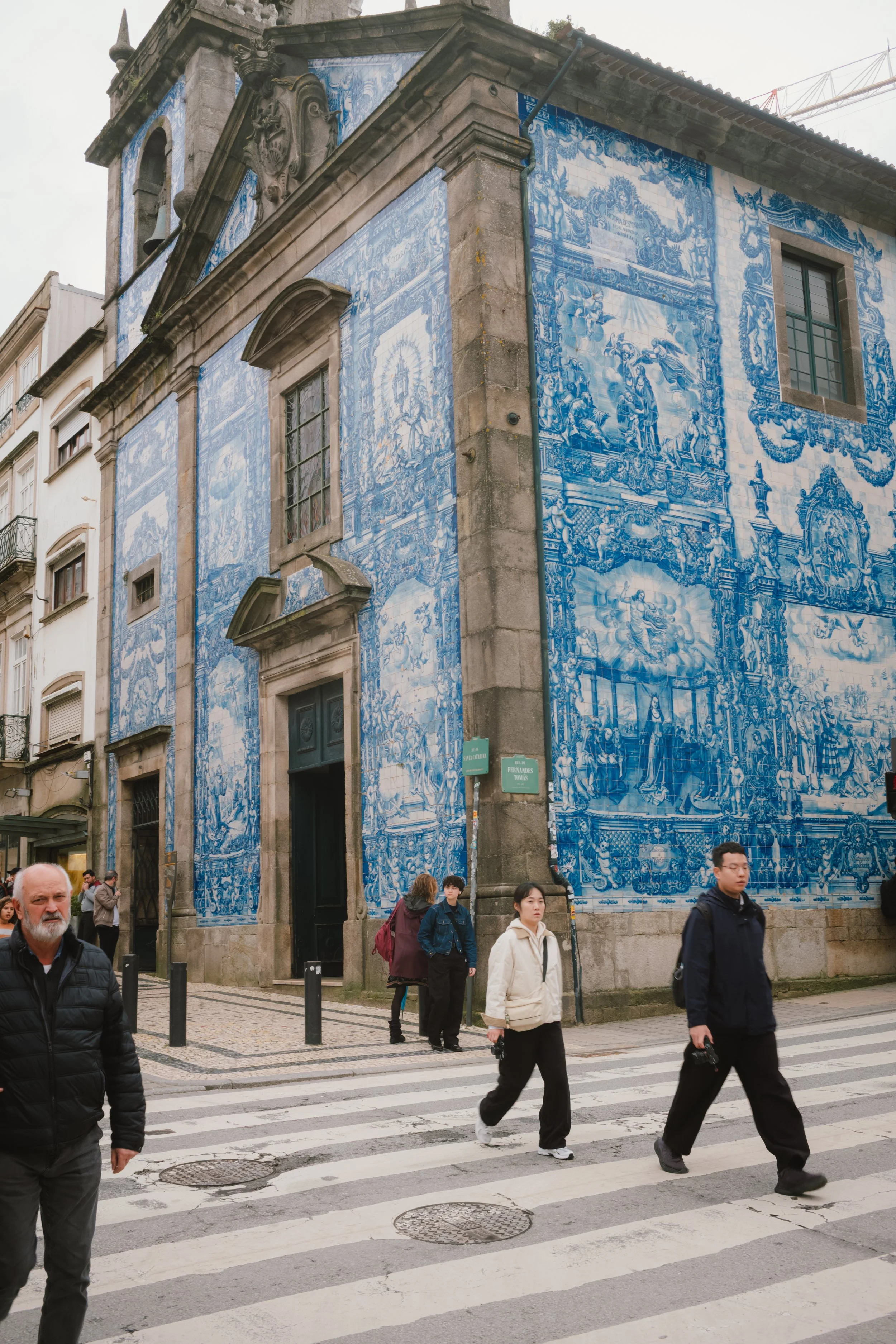A historic building decorated with traditional blue and white azulejo tiles featuring intricate scenes, located at a street corner with pedestrians crossing a zebra-striped crosswalk.