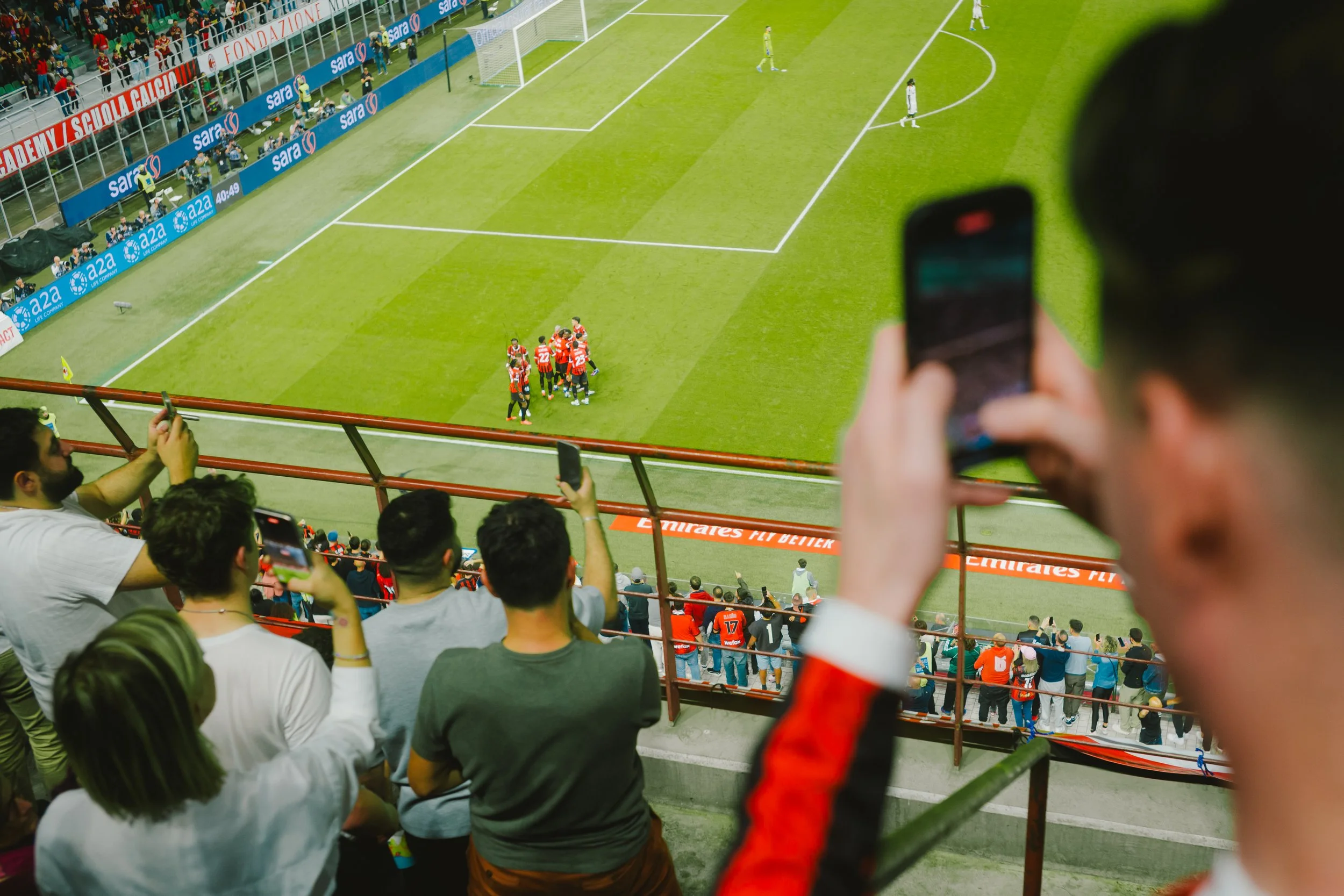 Spectators in the stands watching a soccer game, with some using their phones to take pictures or record. On the field, a group of soccer players wearing red uniforms are celebrating near the sideline, while others in the background stand on the fiel
