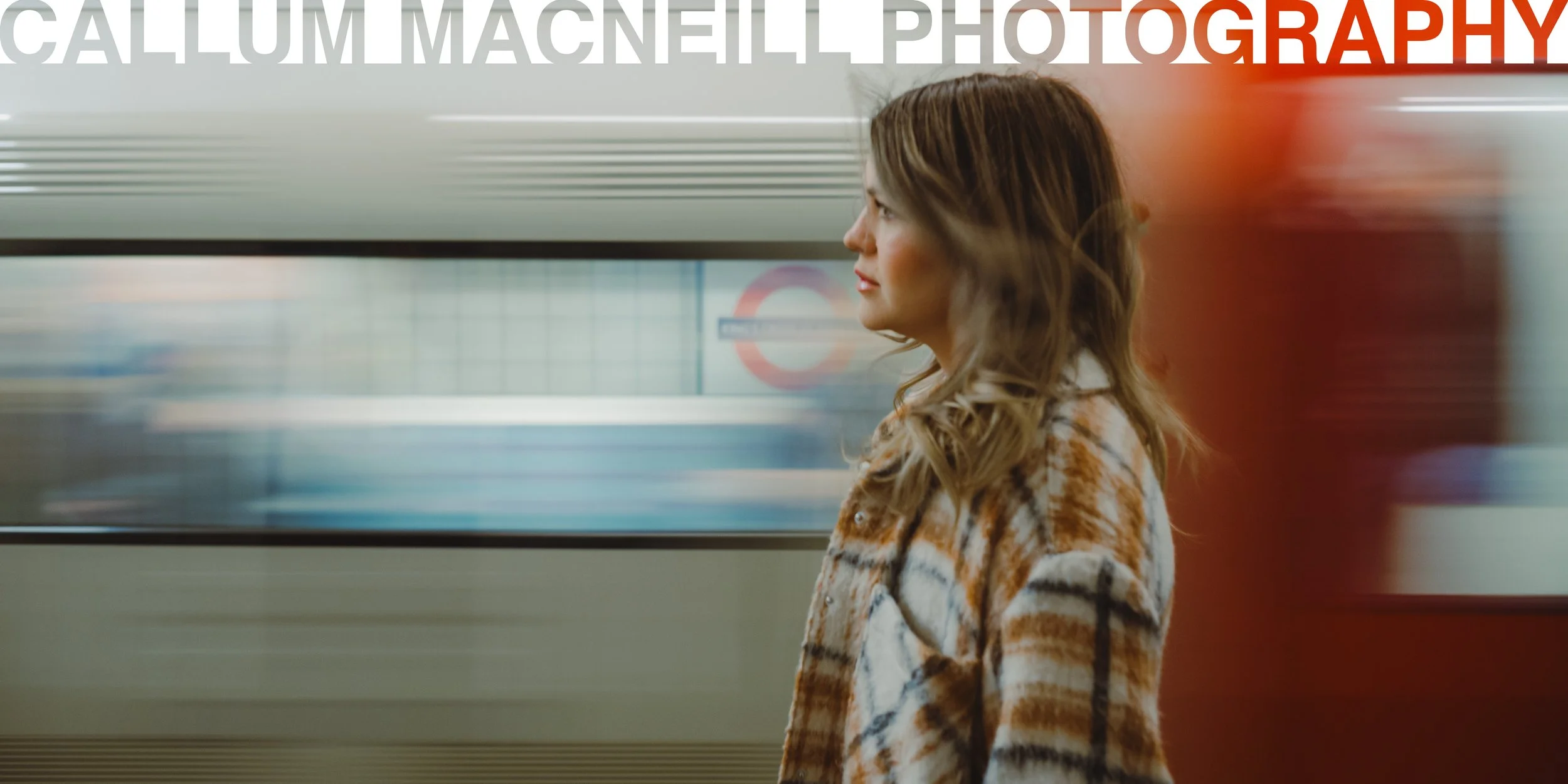 A woman with long wavy hair stands in profile on a subway platform, wearing a patterned coat, with the subway train in motion behind her.
