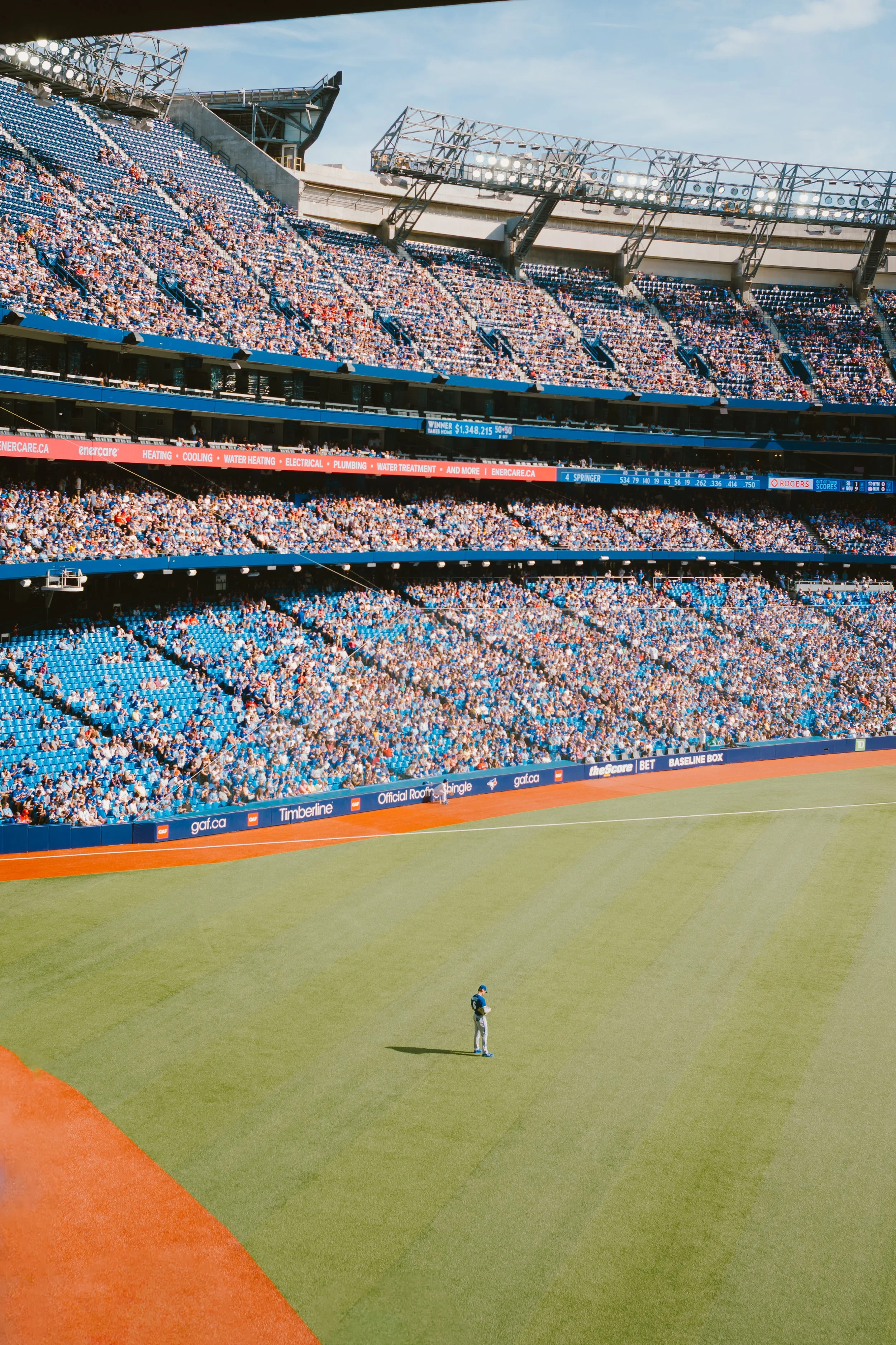 A baseball stadium filled with spectators, with a player standing alone on the field. The outfield has neatly maintained grass, and the infield shows the dirt. There are digital scoreboards and advertisements around the stadium.