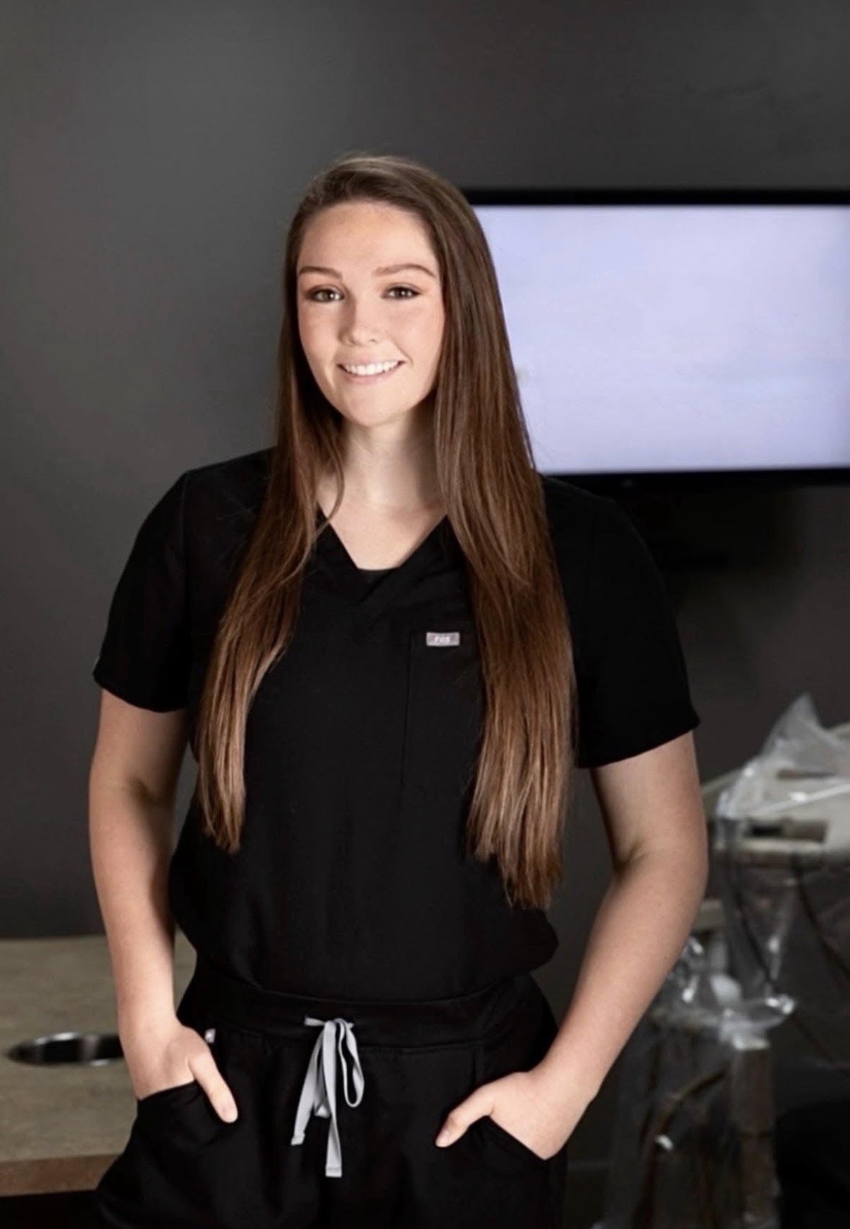 A young woman with long brown hair, smiling, wearing a black uniform, standing inside a room with a gray wall and a television in the background.