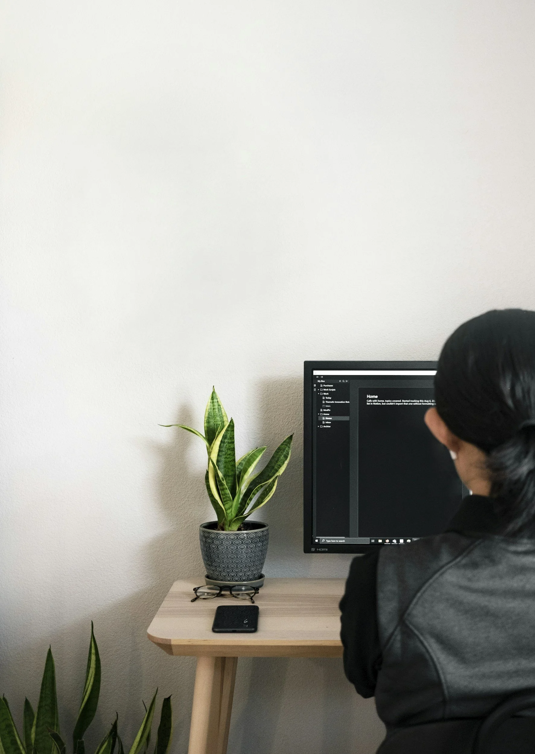 A person with black hair wearing a black and gray jacket sitting at a desk with a computer monitor, a potted snake plant, and a black smartphone on the desk, in front of a white wall.