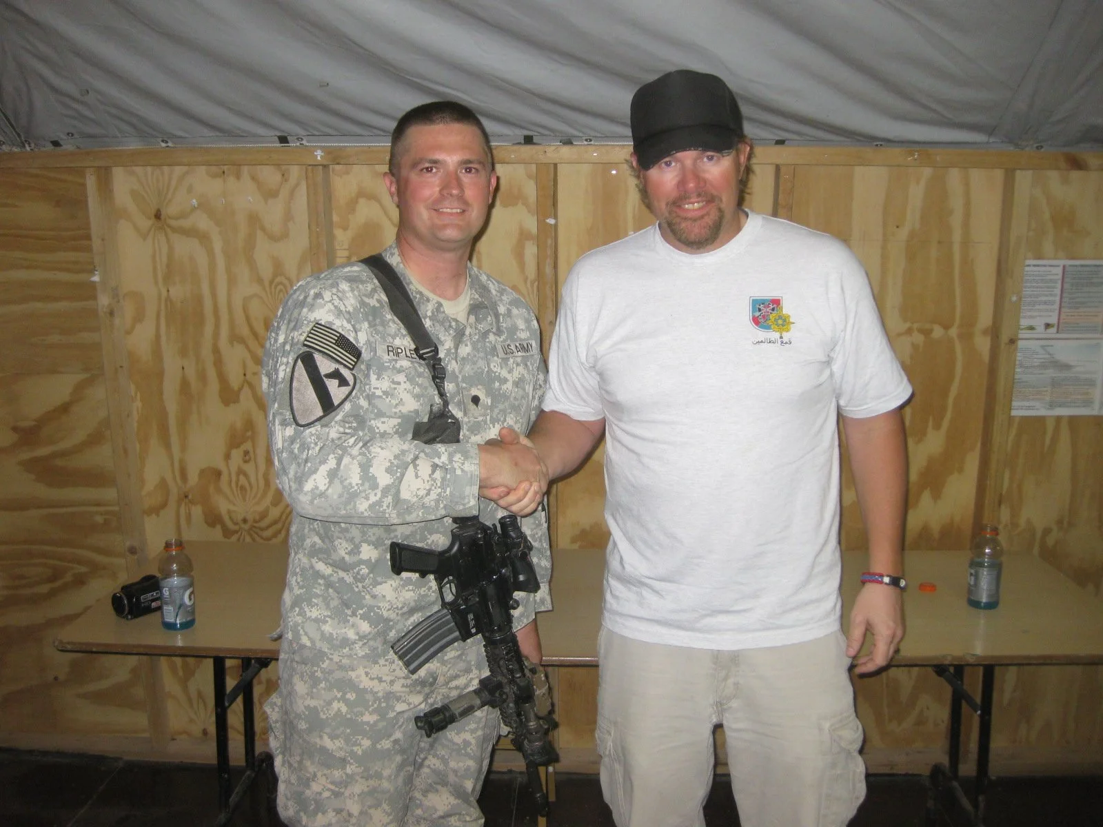 A man in military uniform shaking hands with a man in a white T-shirt inside a wooden structure.