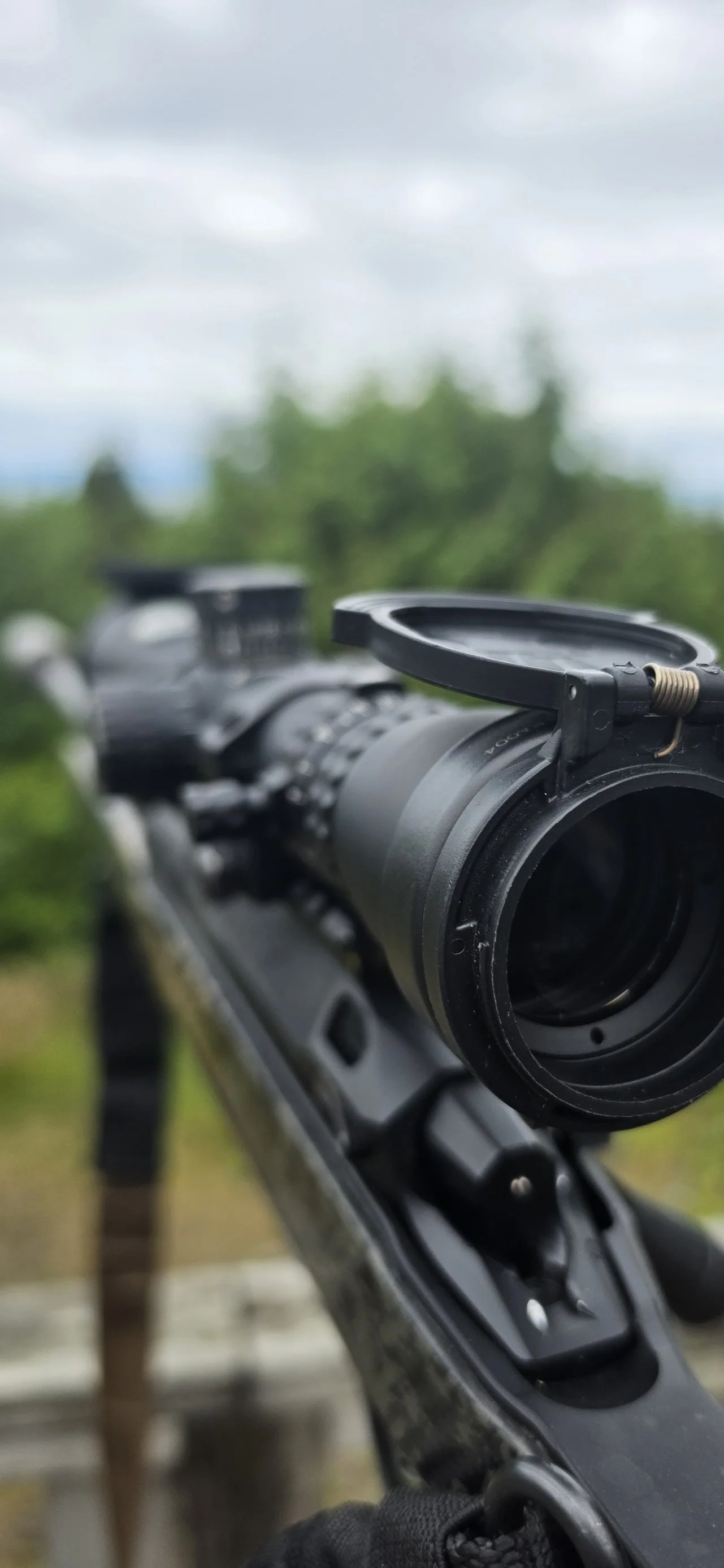 Close-up of a riflescope mounted on a rifle outdoors, with a blurred background of trees and a cloudy sky.