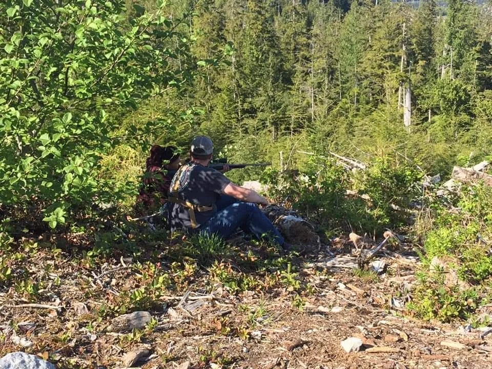 Person sitting in a forested area aiming a rifle with a scope, surrounded by bushes and trees.