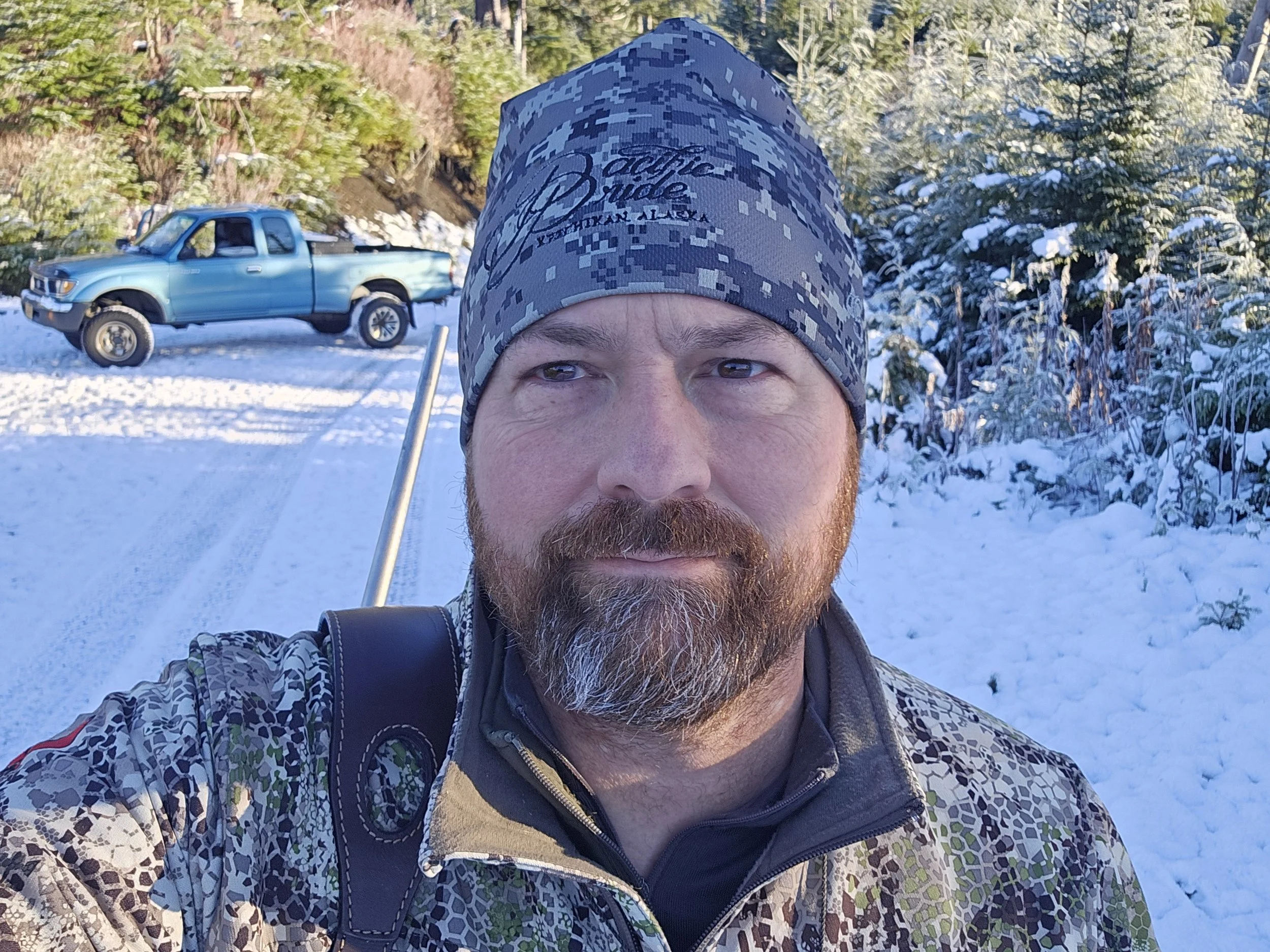 A man with a beard wearing a camouflage patterned jacket and a blue digital camouflage beanie taking a selfie in a snowy forest with a light blue pickup truck in the background.
