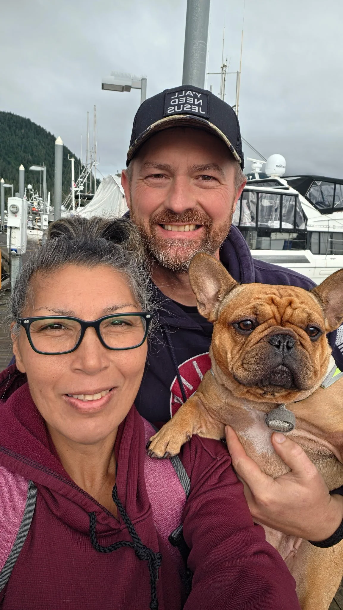 A smiling man and woman taking a selfie with a French Bulldog at a marina, with boats and cloudy sky in the background.