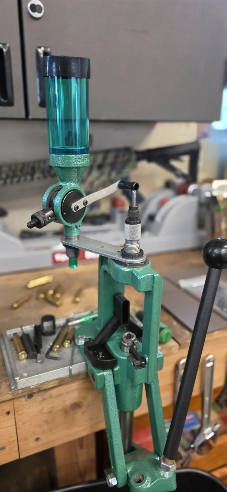 A brass reloading press used for resizing and priming cartridge cases, with a powder measure attached on top, situated on a wooden workbench surrounded by brass casings and reloading tools.