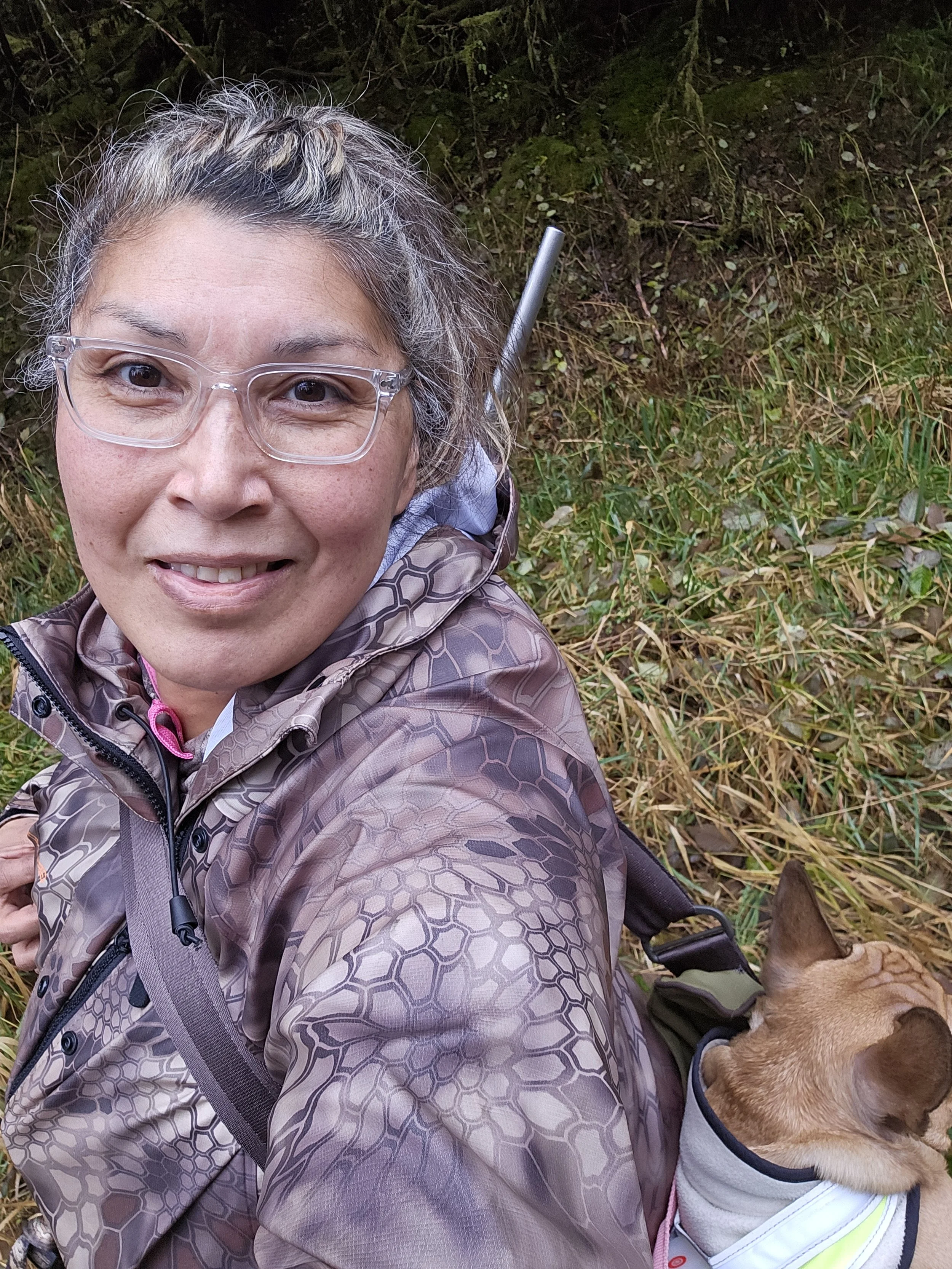 A woman with glasses and gray hair taking a selfie outdoors in a forested area with a small dog in a white hoodie.