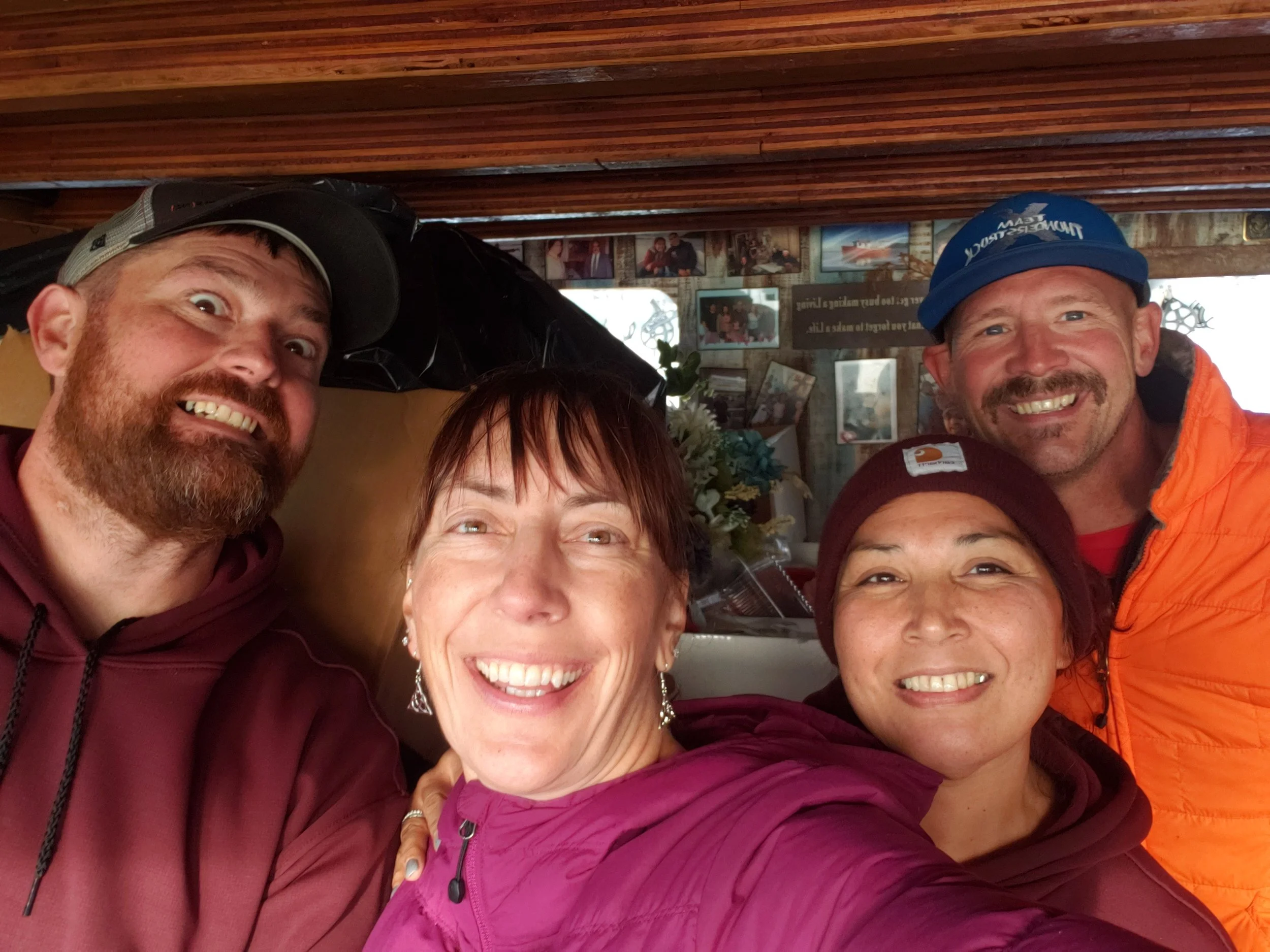 Five smiling people wearing outdoor gear, close together in a rustic indoor setting with wood-paneled ceiling and framed pictures on the wall.