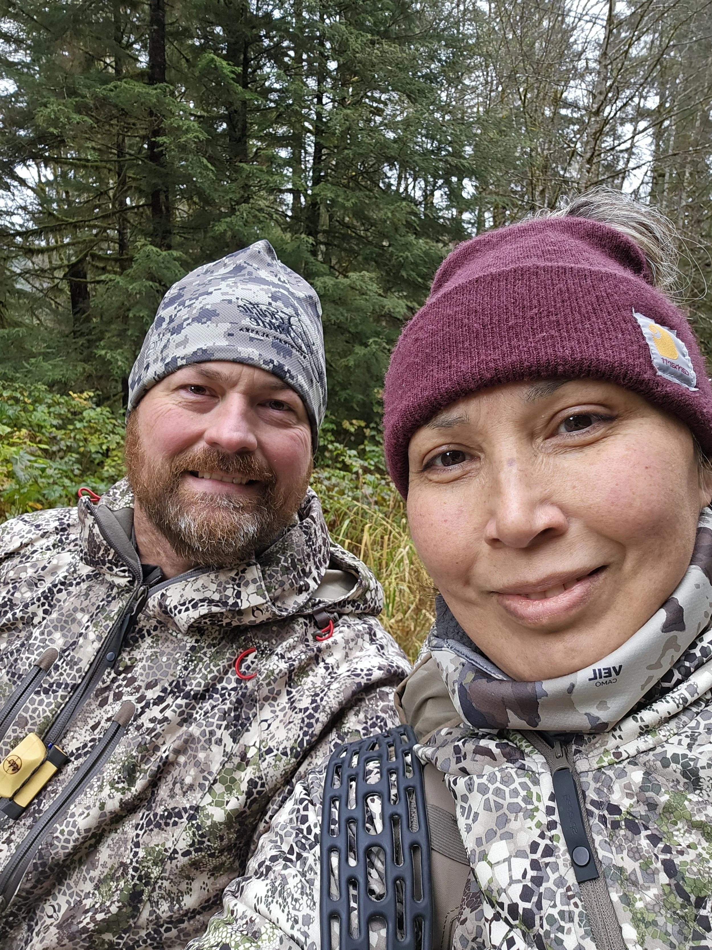 A man and a woman outdoors in a forested area, wearing camouflage jackets and beanies. The man has a gray and white beanie and a beard, and the woman has a maroon beanie. Both are smiling and close together.