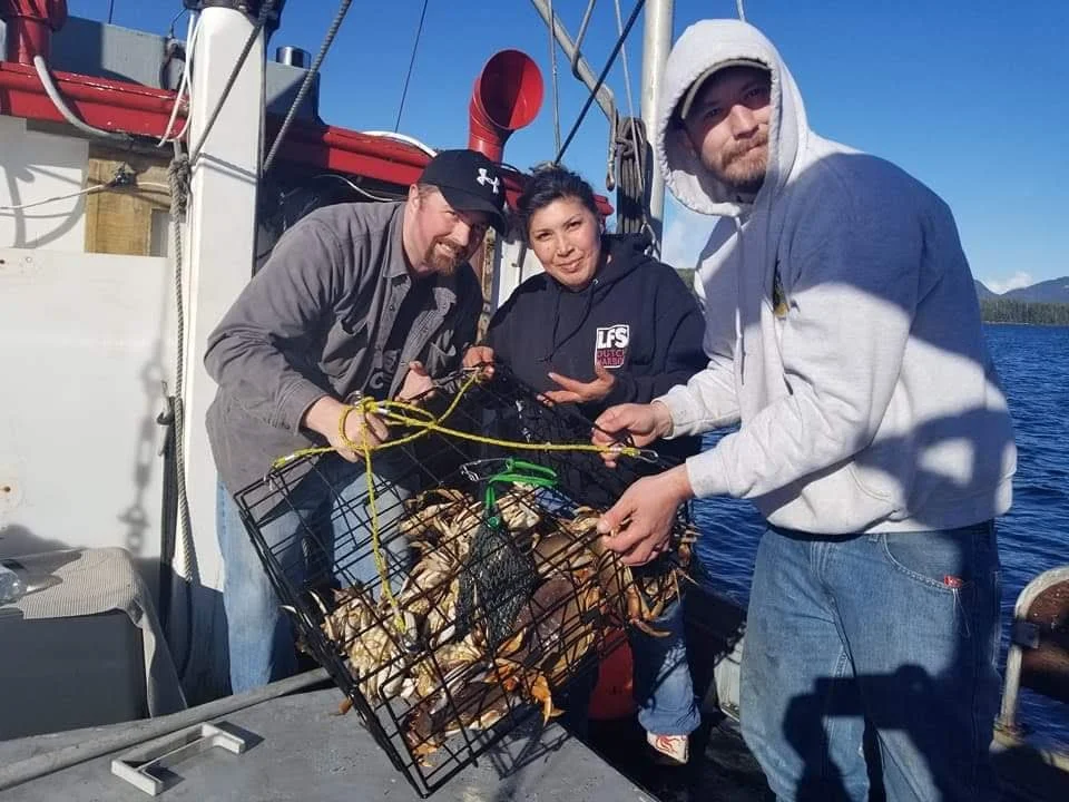 Three people on a boat holding a crab trap with crabs inside, with a blue sky and water in the background.