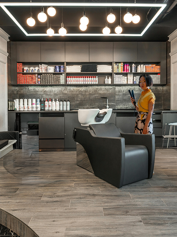 Interior of a modern hair salon with black leather chairs, a hair washing station, and shelves stocked with hair products. A woman in a yellow top and patterned skirt stands near the wall, holding a phone.