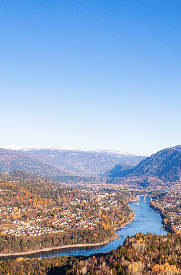 A panoramic view of Columbia River winding through a valley surrounded by mountains with snow-capped peaks in the distance, and a small city of Castlegar BC along the riverbanks.