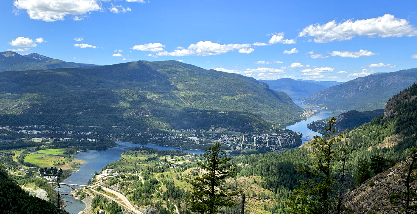 A panoramic view of a river winding through a green valley surrounded by mountains, under a partly cloudy blue sky.
