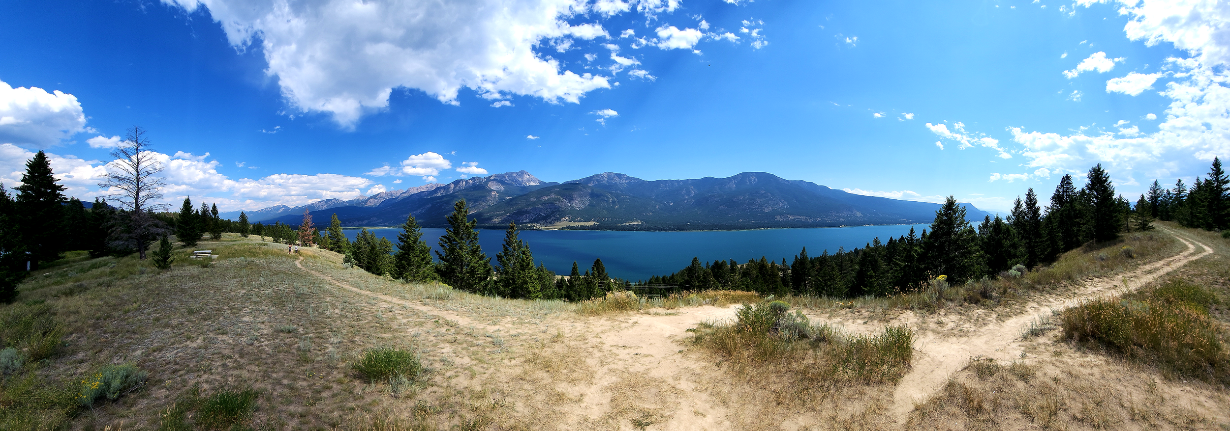 A panoramic view of a lake surrounded by mountains under a blue sky with scattered clouds. In the foreground, there is a dirt trail and a grassy area with scattered trees including some evergreens.