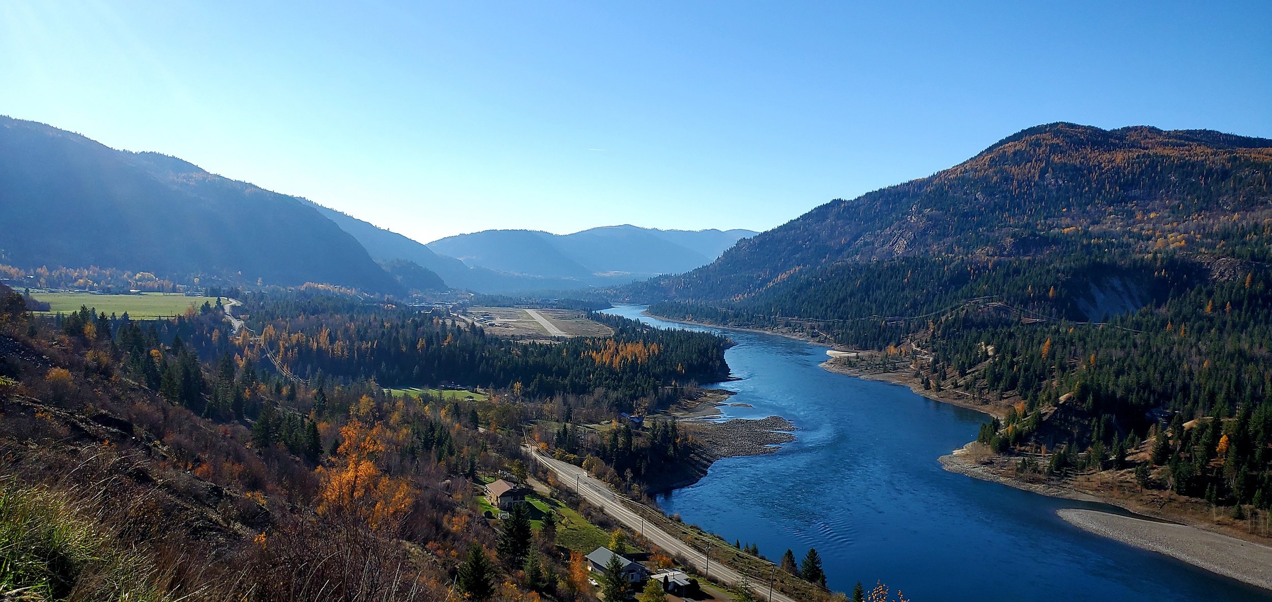 A scenic view of a river winding through a valley with forested hills and mountains on both sides, under a clear blue sky.