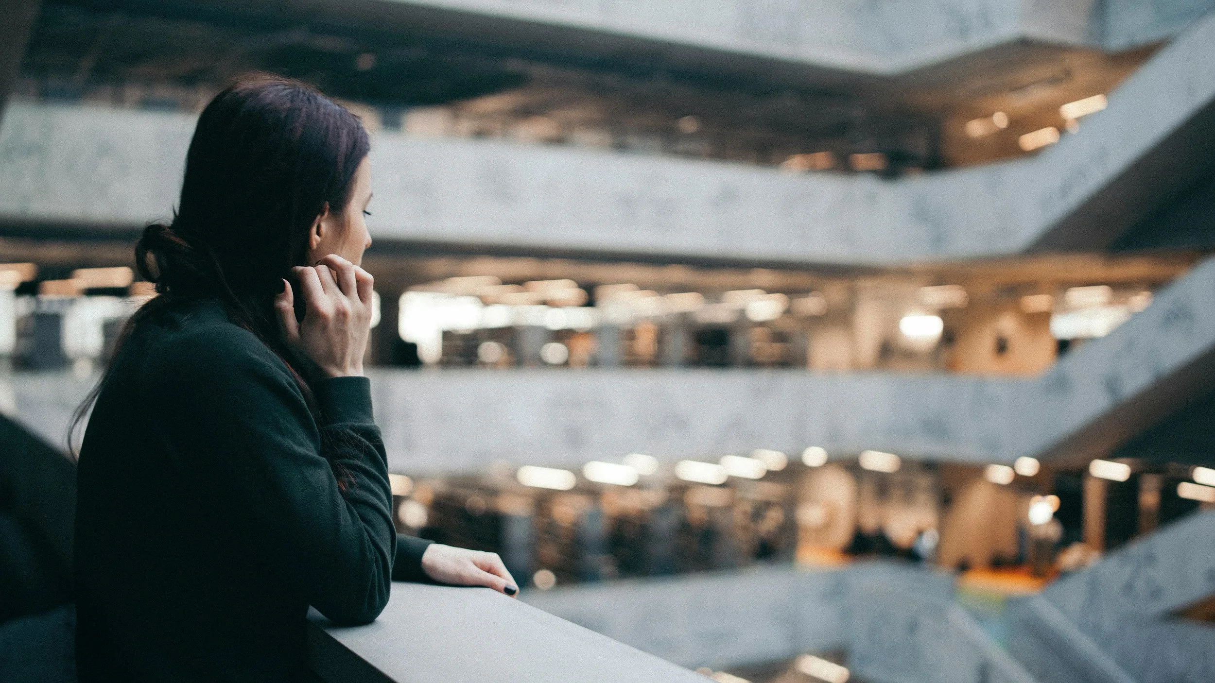 A woman with dark hair in a ponytail, wearing a black top, is leaning on a railing in an indoor building, looking to the side with her hand near her face. The background shows multi-level parking garage with soft lighting and blurred structural elements.
