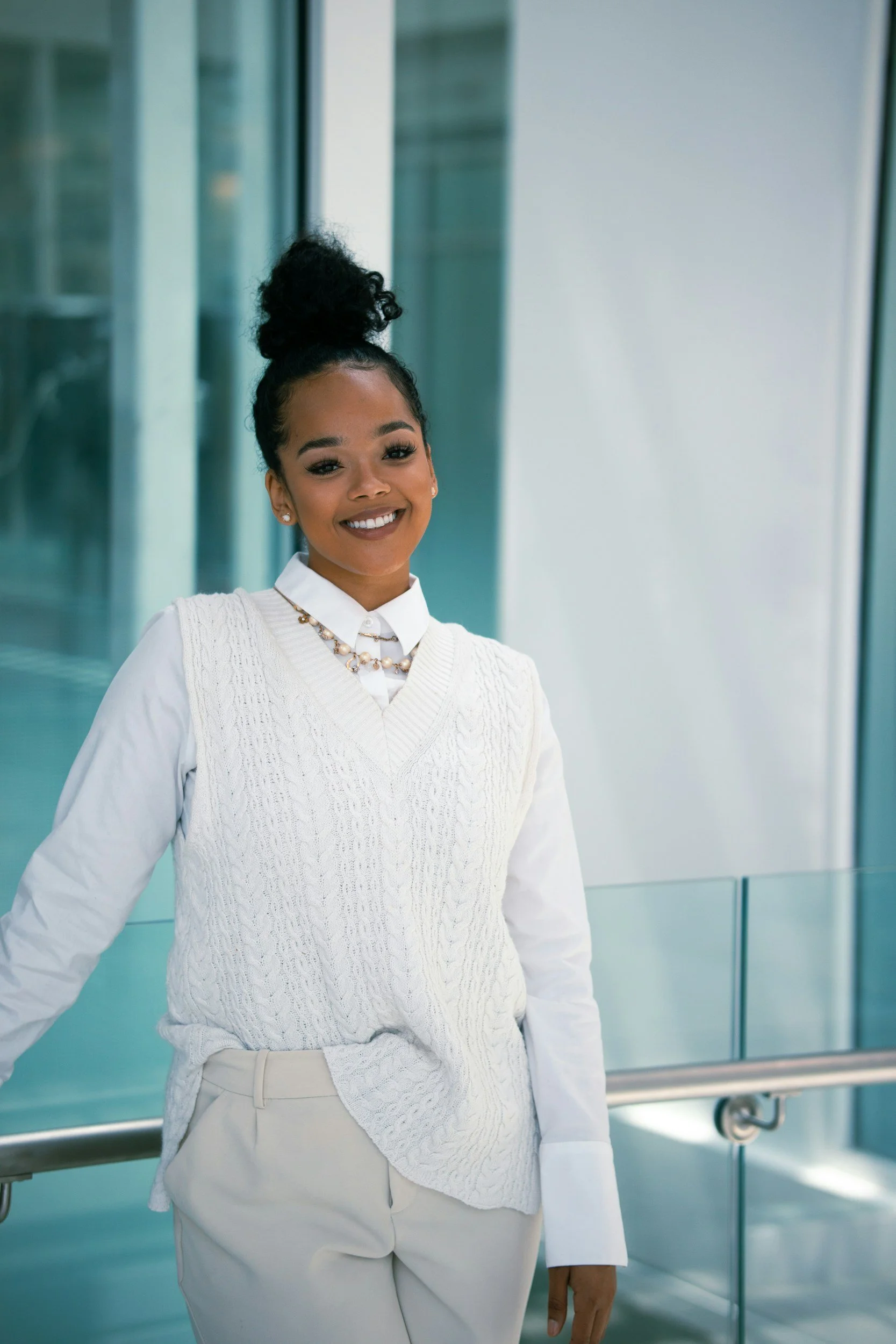 A young woman smiling and standing in front of glass windows, wearing a white cable knit vest over a white shirt, accessorized with pearl jewelry, and beige pants.
