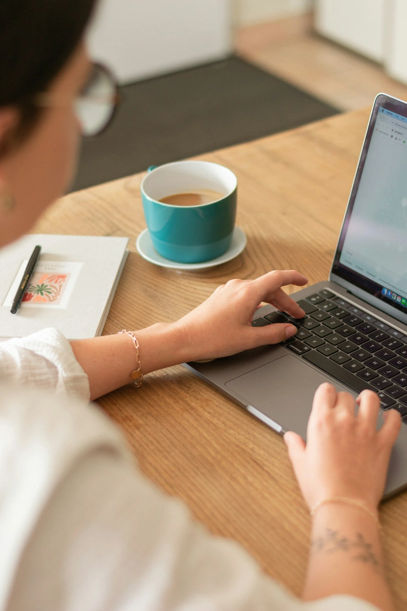 A person using a laptop at a wooden table with a cup of coffee, a notebook, and a pen, in a cozy indoor setting.
