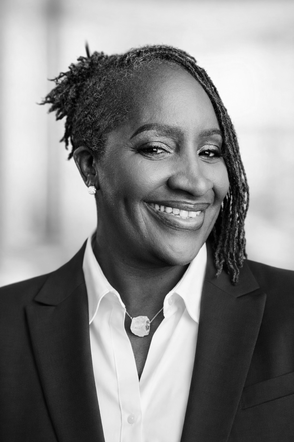 Black and white portrait of a smiling professional woman wearing a suit, white shirt, necklace, and earrings, with styled dreadlocks.