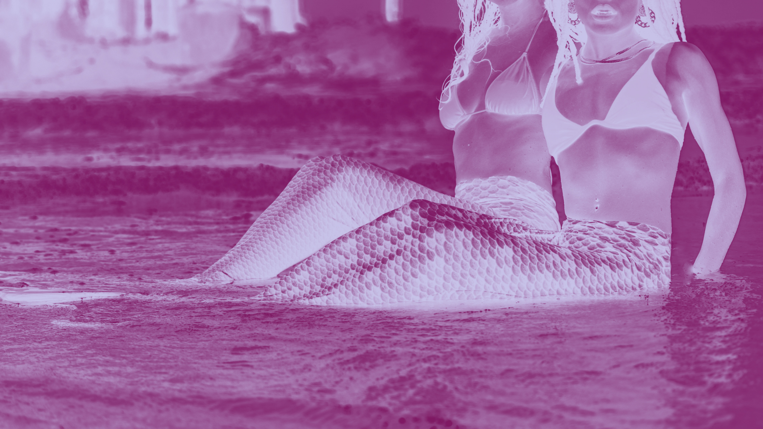A woman with a mermaid tail sitting in water at the beach, wearing a bikini top.