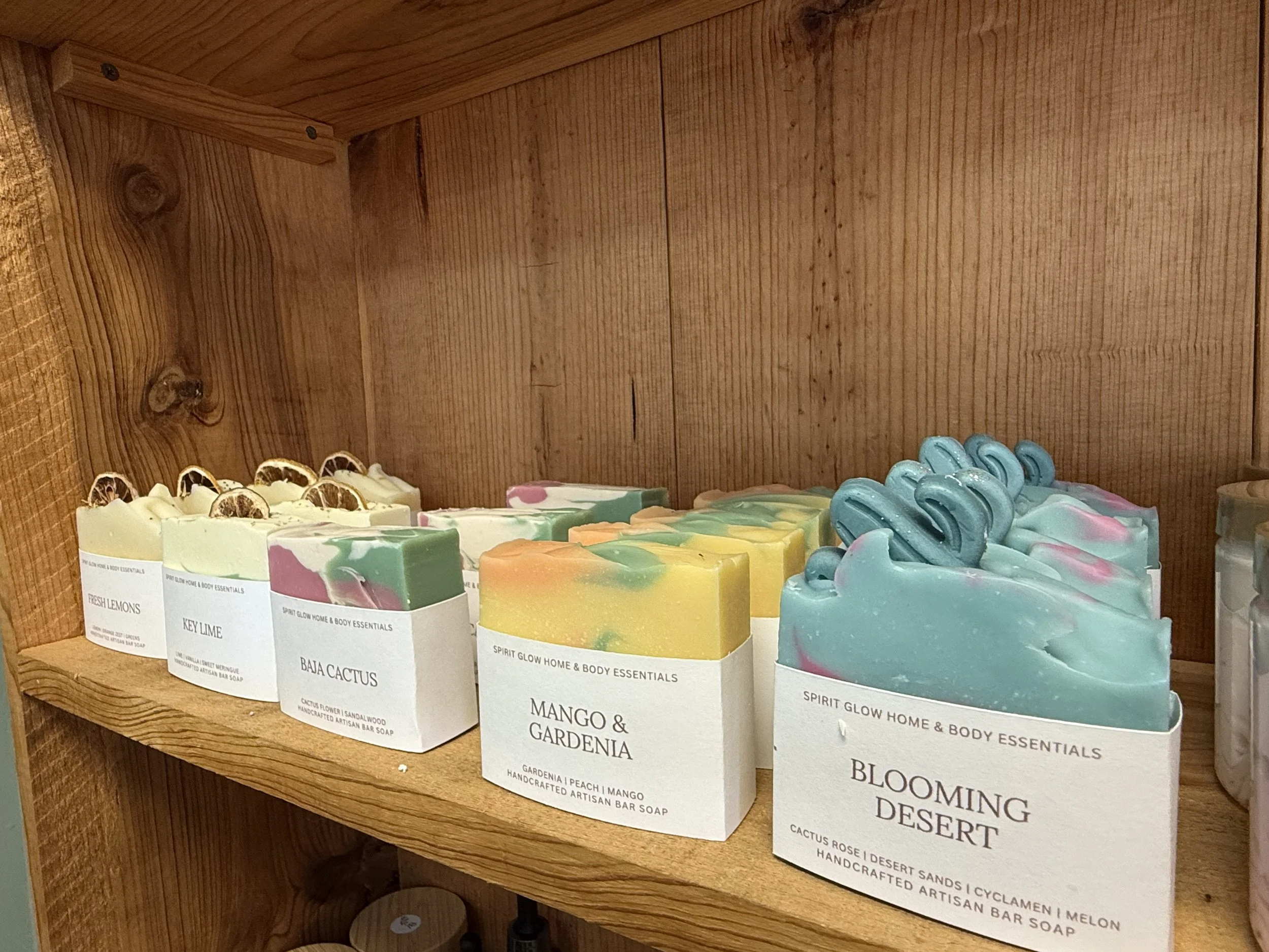 Various colorful handmade artisanal bar soaps on a wooden shelf, with labels including scents like fresh lemons, key lime, mango & gardenia, blooming desert, and cactus rose in desert sands, in a store or display setting.