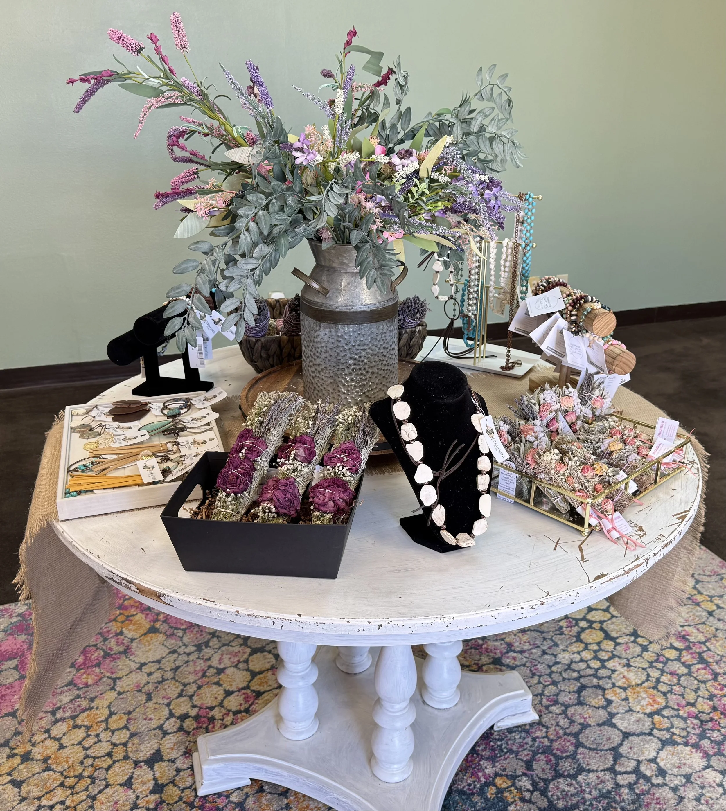 A round white distressed wood table displaying jewelry including necklaces, earrings, bracelets, and hair accessories, with a large floral arrangement at the center in a rustic metal pitcher.