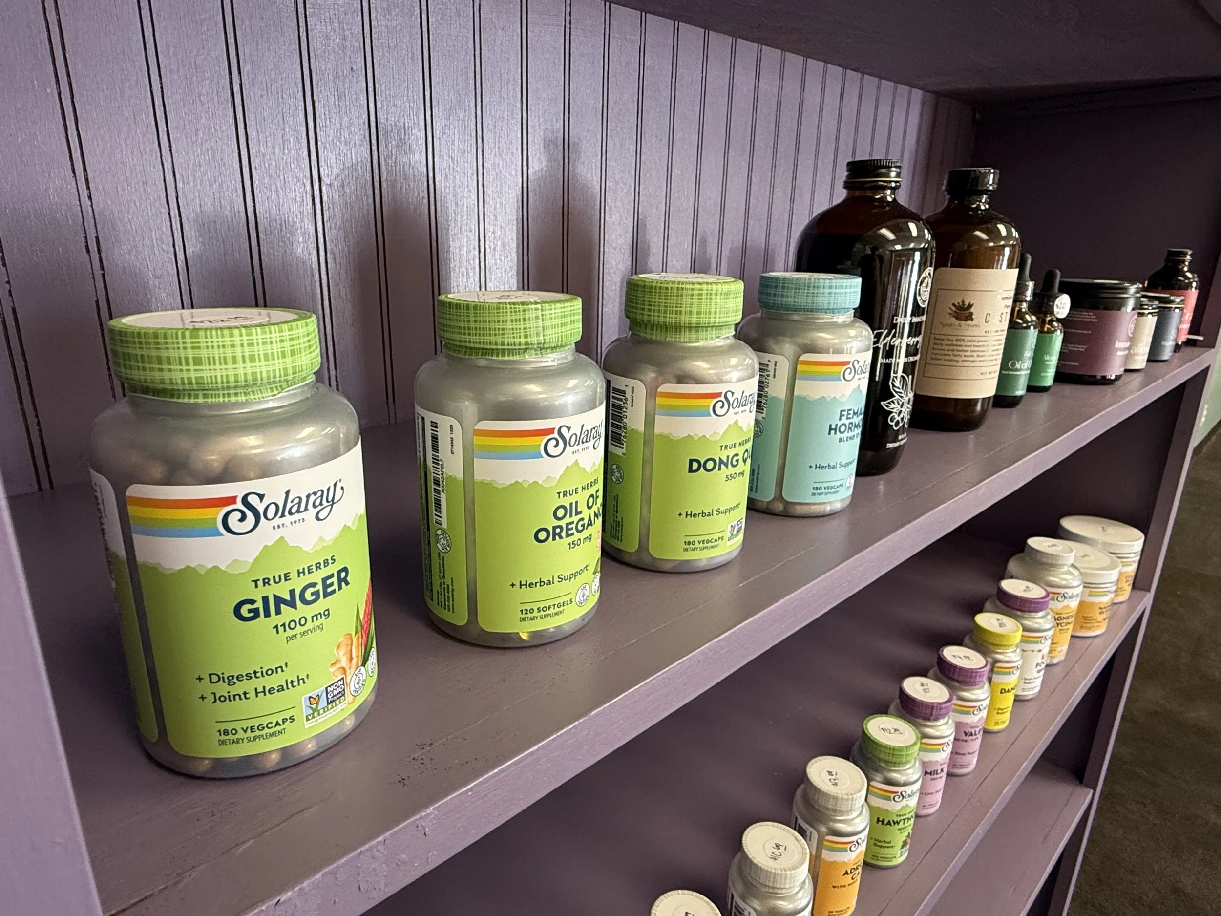 A purple shelf holding various bottles of herbal supplements and vitamins. The bottles are mainly green and brown, with some smaller bottles and capsules visible.